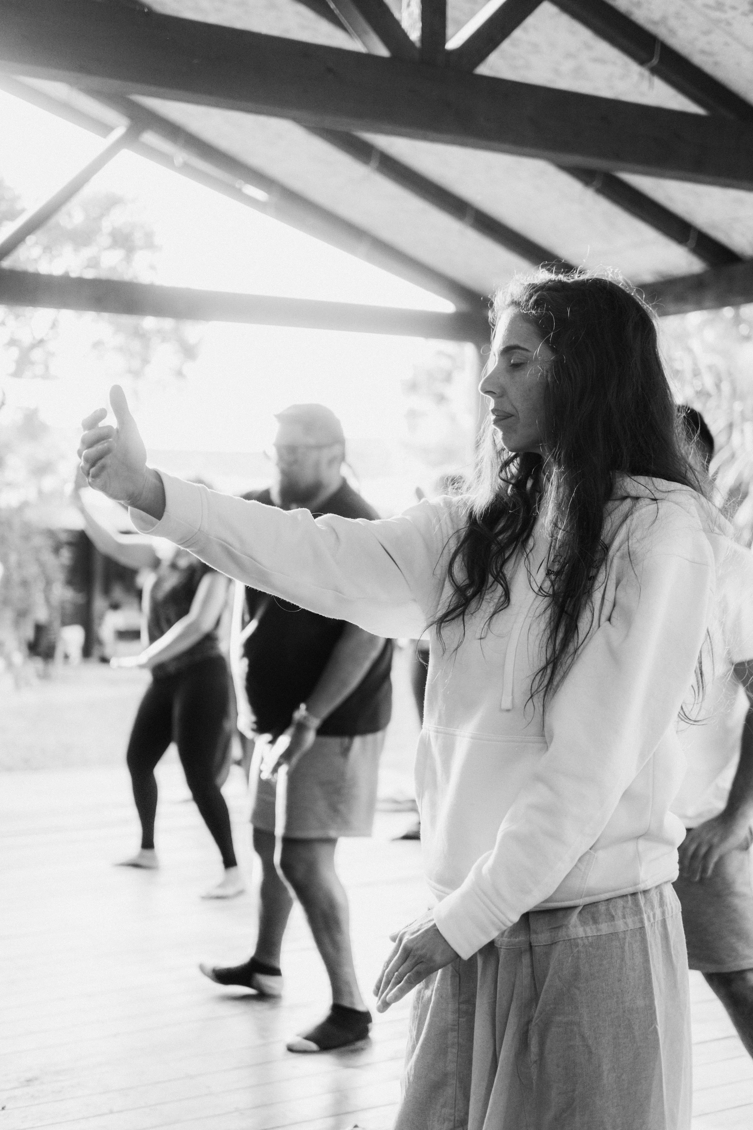 Mujer con cabello largo y rubio, con los ojos cerrados, practicando meditación en un lugar con estructura de madera, junto a otras personas que también parecen practicar alguna actividad en un espacio cubierto, en un entorno natural.
