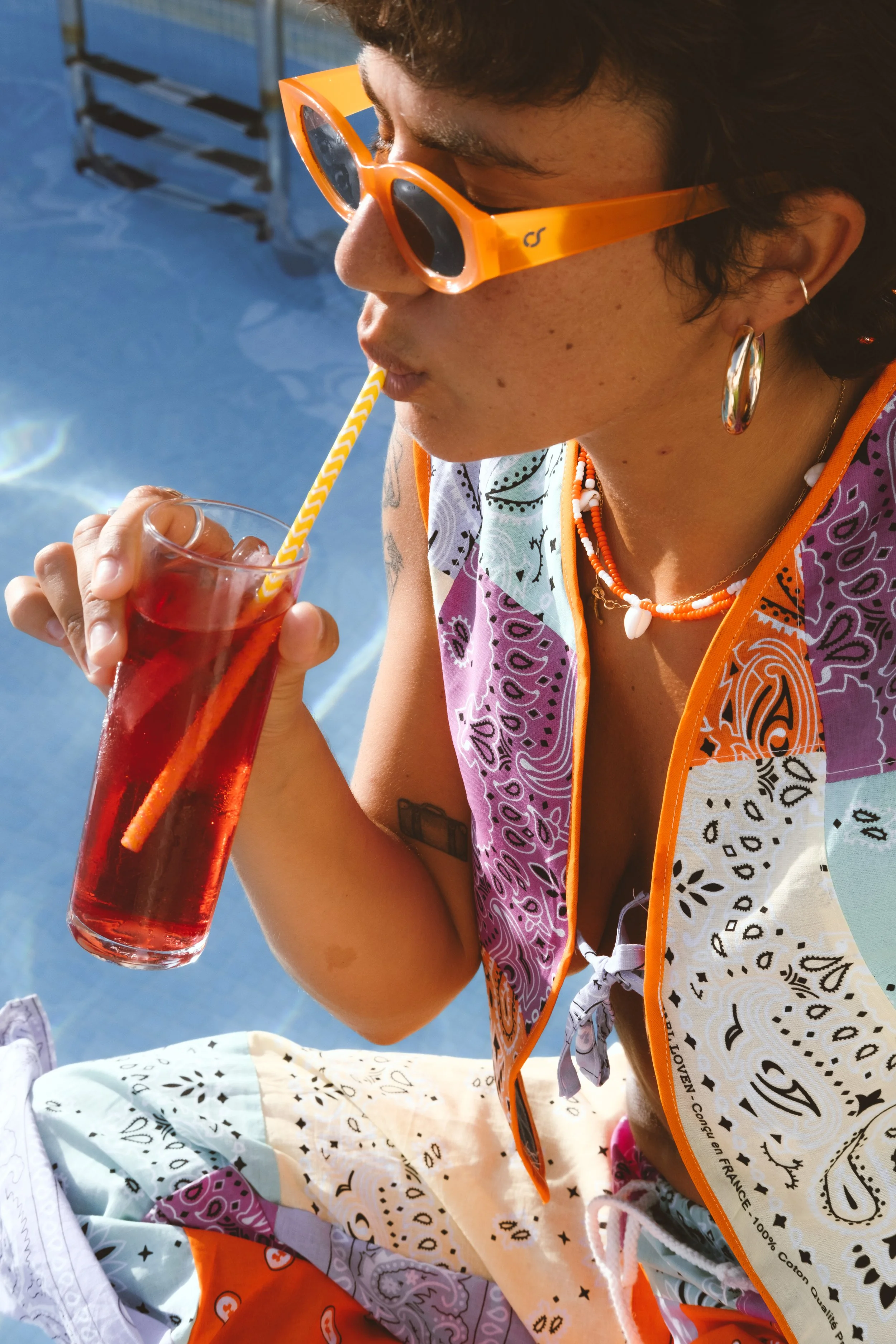 Mujer usando gafas de sol anaranjadas, bebiendo una bebida roja a través de una pajilla amarilla con blanco en un ambiente al aire libre.