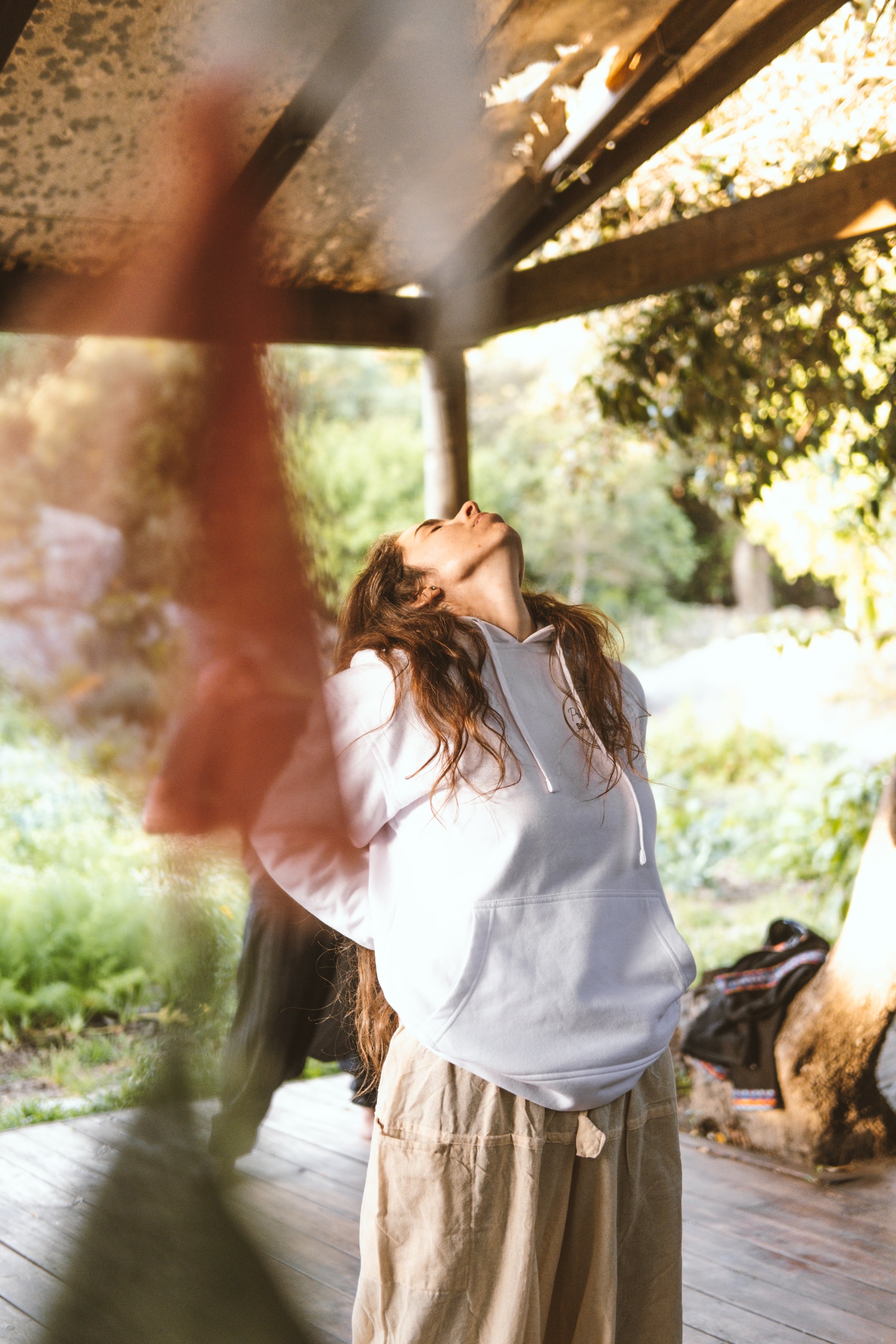 Una mujer con cabello rizado y largo, vestida con sudadera gris y pantalones beige, está de pie viendo hacia arriba en un entorno natural con árboles y una estructura de madera.
