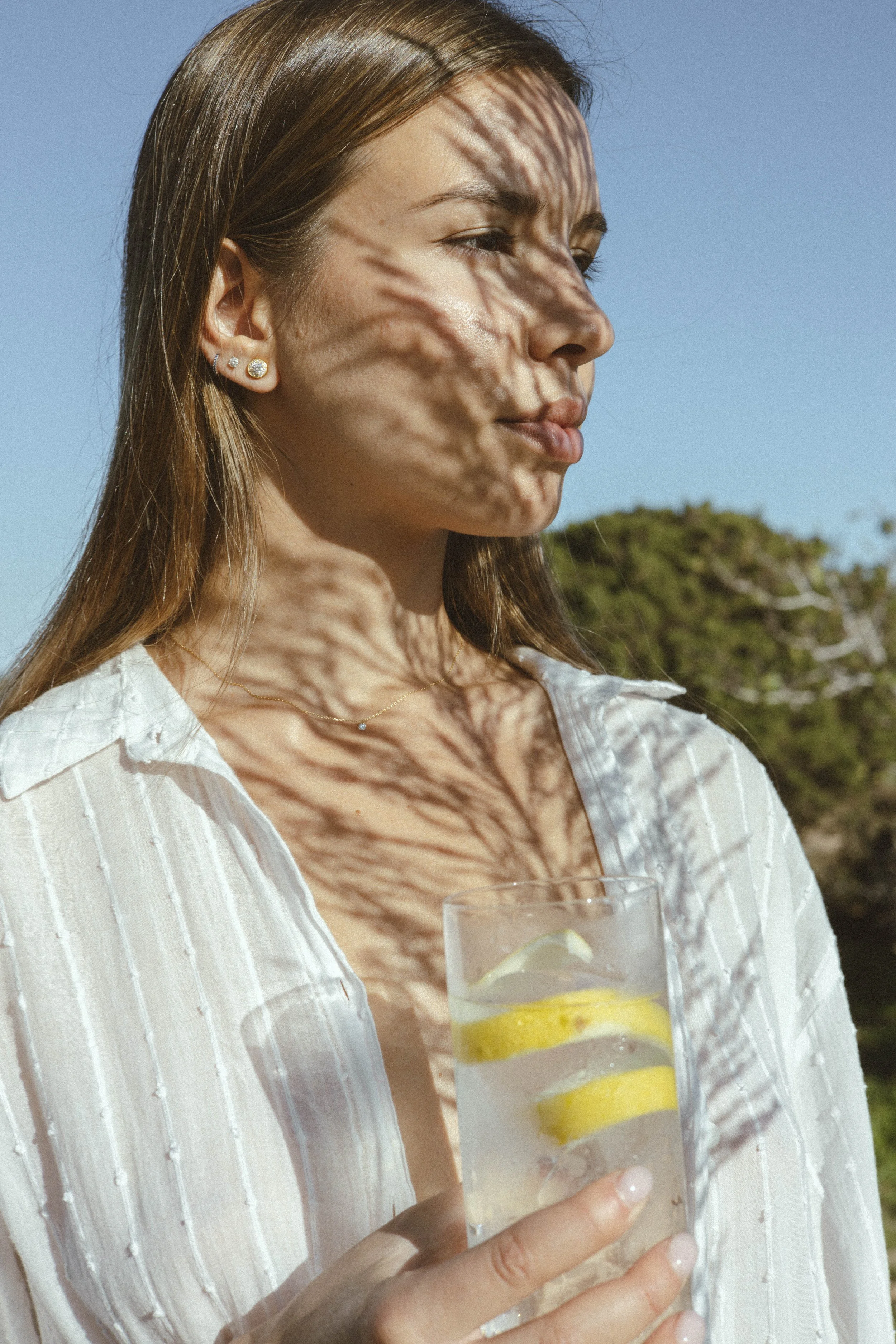 Mujer joven con cabello castaño claro, vistiendo blusa blanca de rayas y pendientes de diamantes, sosteniendo un vaso con agua y rodajas de limón, en un entorno exterior con cielo azul y árboles de fondo, sombra de hojas proyectada en su rostro.