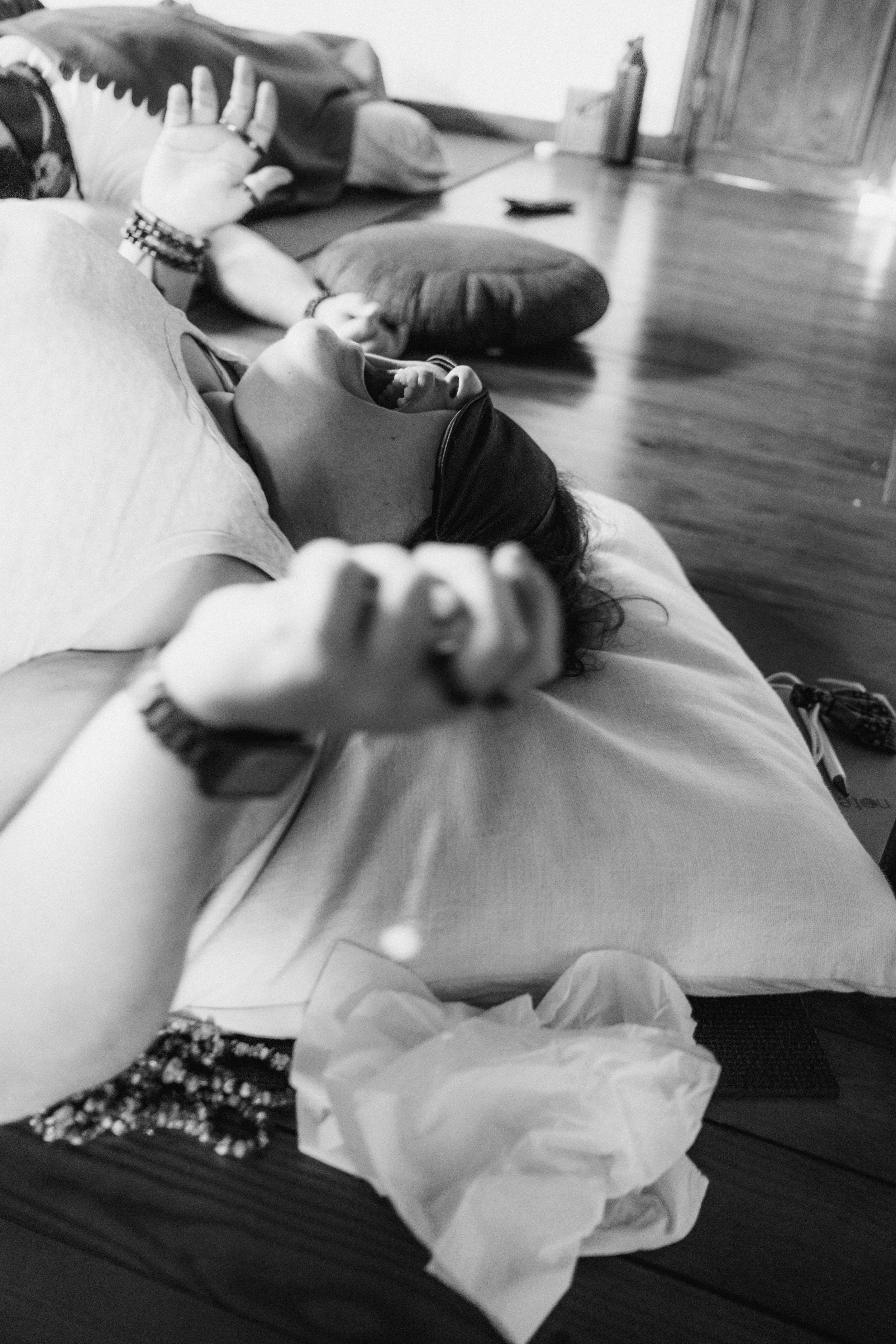 Una persona acostada en una cama, con la boca abierta, parece estar durmiendo o hablando. Tiene pulseras en la muñeca y está vestida con ropa de manga larga. La imagen está en blanco y negro y en el fondo se puede ver una habitación con pisos de made