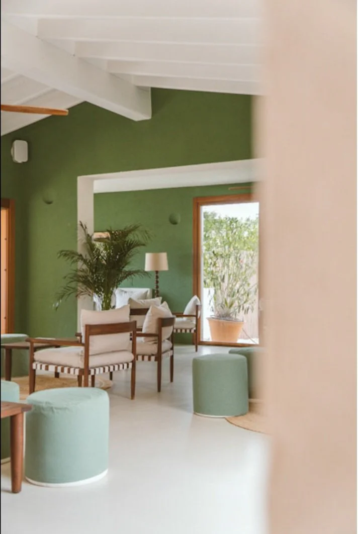 Sala de estar con paredes verdes, pantallas de luz en la pared, plantas y muebles de madera con cojines blancos y verdes, con ventanas grandes que dejan entrar luz natural.
