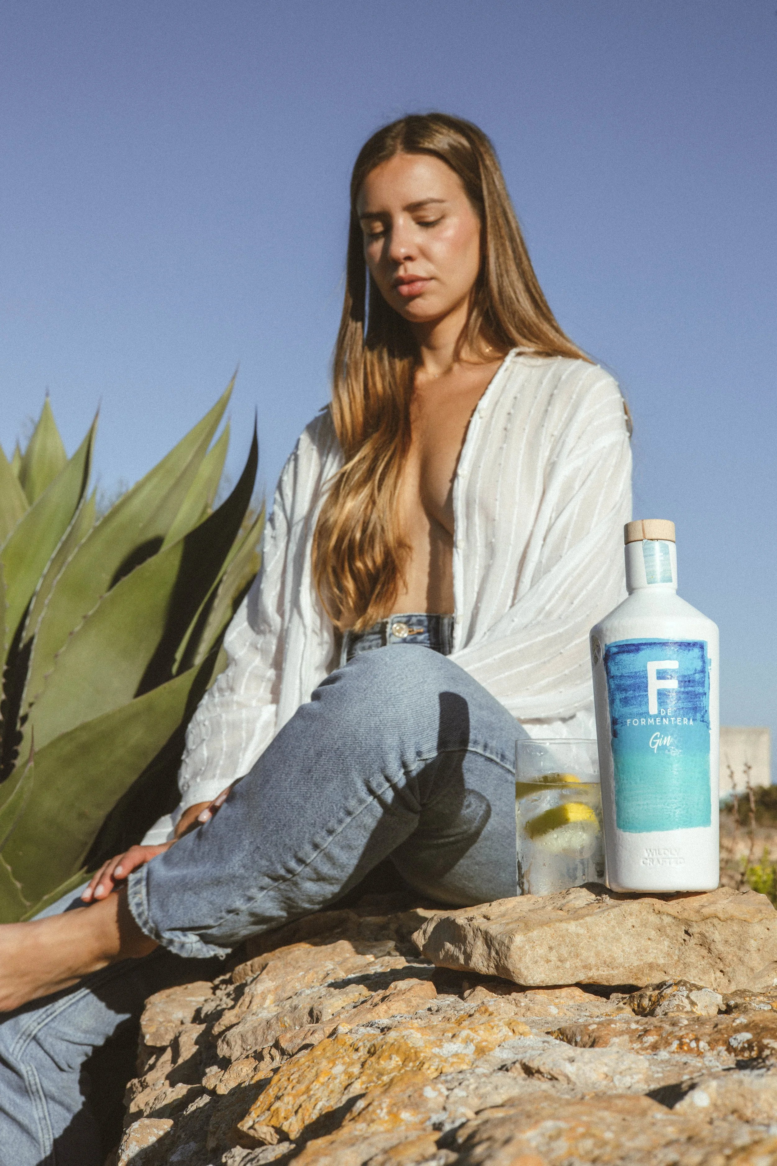 Joven mujer con cabello largo y suelto, vestida con blusa blanca y jeans, sentada sobre rocas en un entorno exterior con plantas y un cielo despejado, disfrutando de un momento de relajación junto a una botella de ginebra y un vaso con hielo y rodajas de limón.