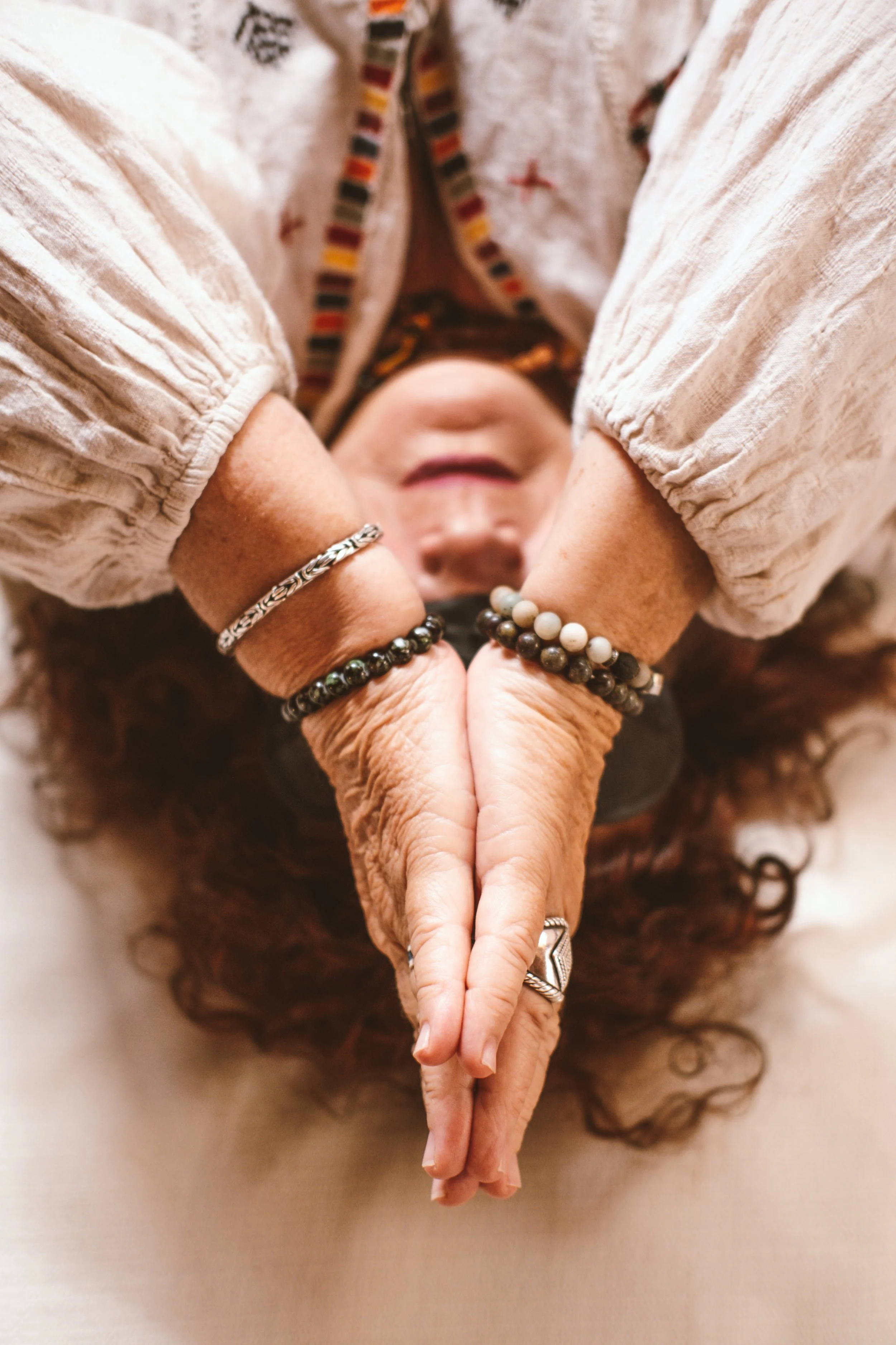 Manos de una persona mayor con varias pulseras y anillos formando una figura de corazón, vista desde abajo, con una expresión de alegría o paz en su rostro.
