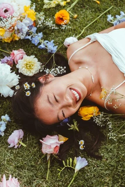 Mujer sonriendo, acostada en césped, rodeada de flores de colores.