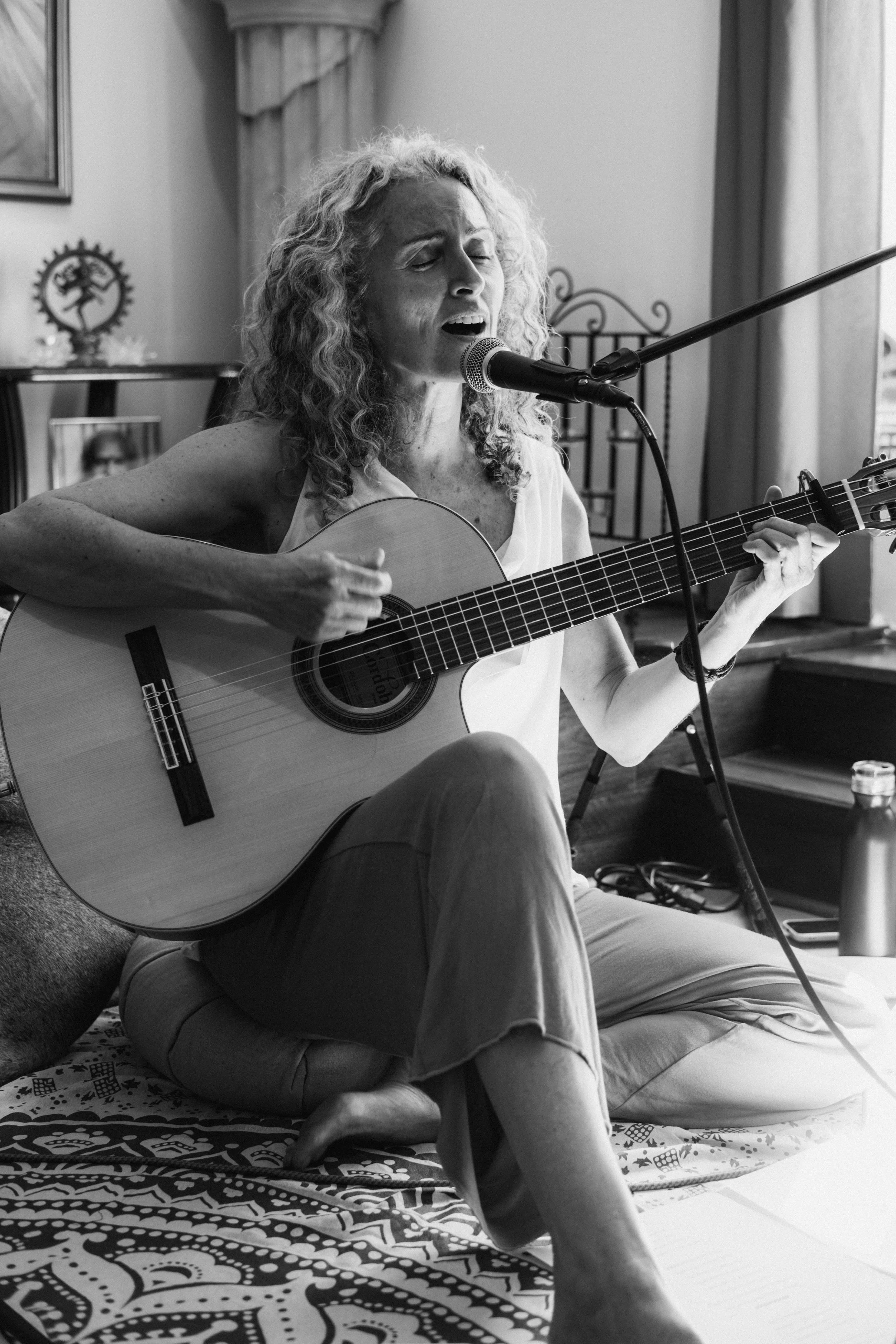 Mujer cantando y tocando guitarra acústica, sentada en el suelo, con micrófono frente a ella, en un ambiente interior.