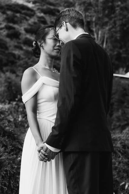 Una pareja en vestido de boda y traje formal en una ceremonia al aire libre, sosteniéndose de las manos y acercándose con las frente juntas.