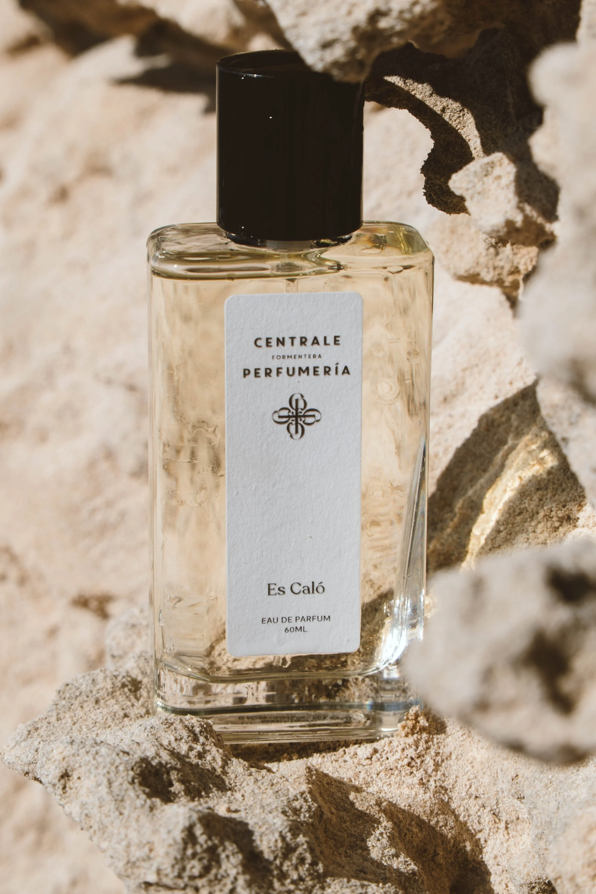 Frasco de perfume transparente con tapa negra, colocado sobre arena y rocas.