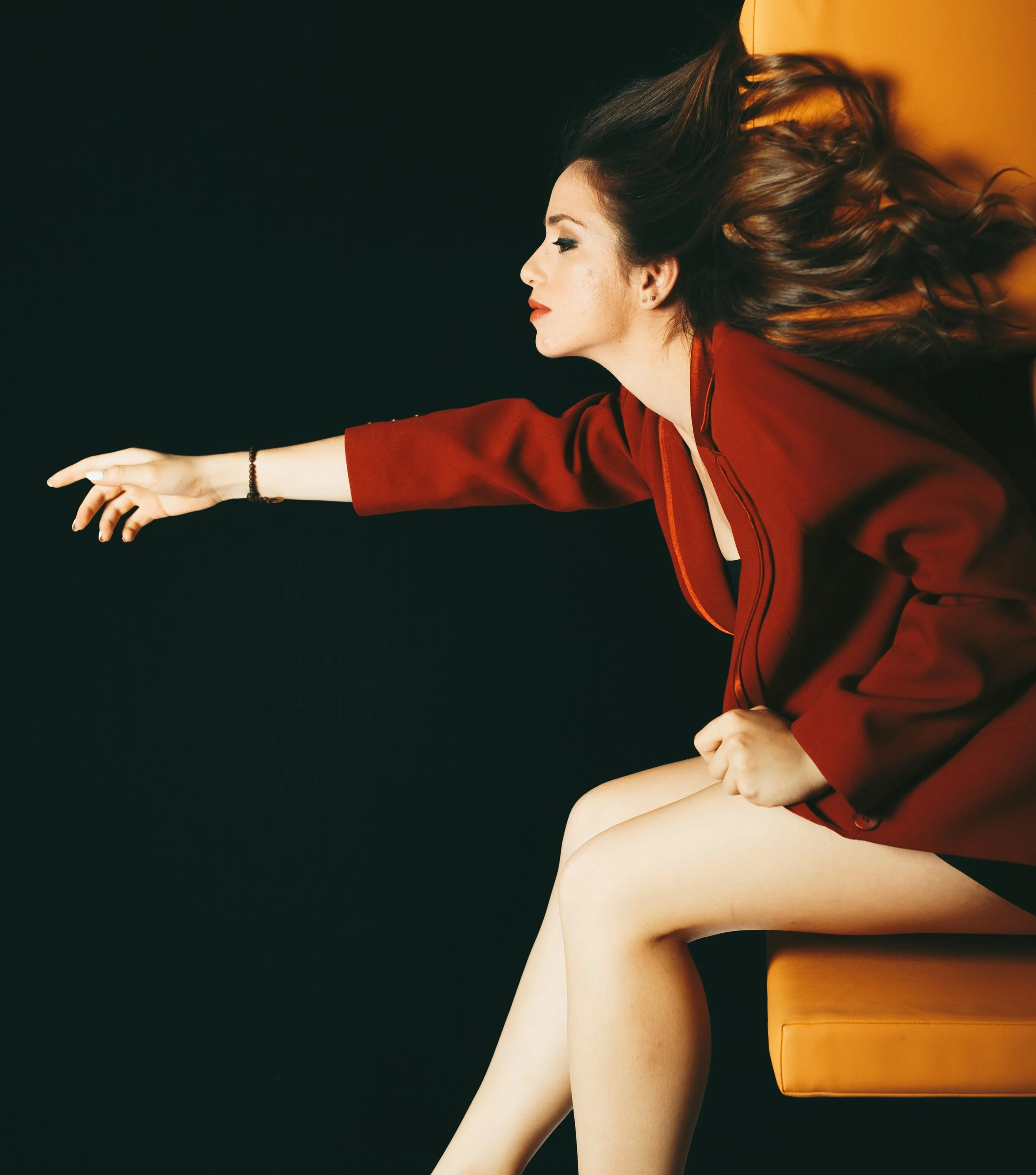 Mujer con cabello rizado que Extiende la mano hacia la izquierda, sentada junto a un mueble naranja, vestida con blusa roja, con fondo oscuro.