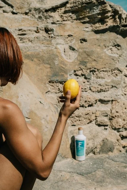 Mujer con cabello castaño sosteniendo un limón amarillo frente a una pared de piedra, con una botella de protección solar en una roca.