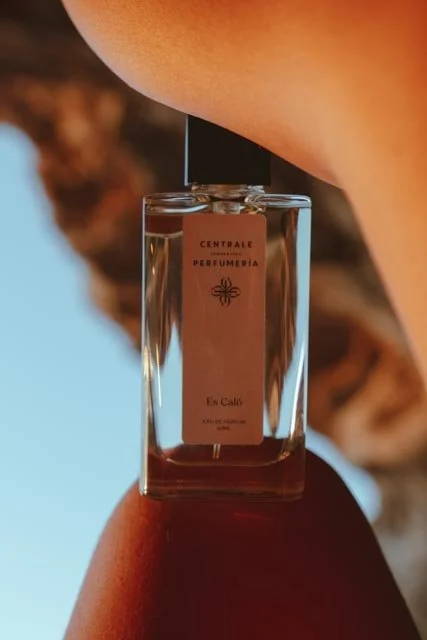Frasco de perfume de perfume, etiquetado con 'Centrale Perfumería' y 'Es Caló', sostenido por una mano sobre la piel, con fondo de cielo y roca.