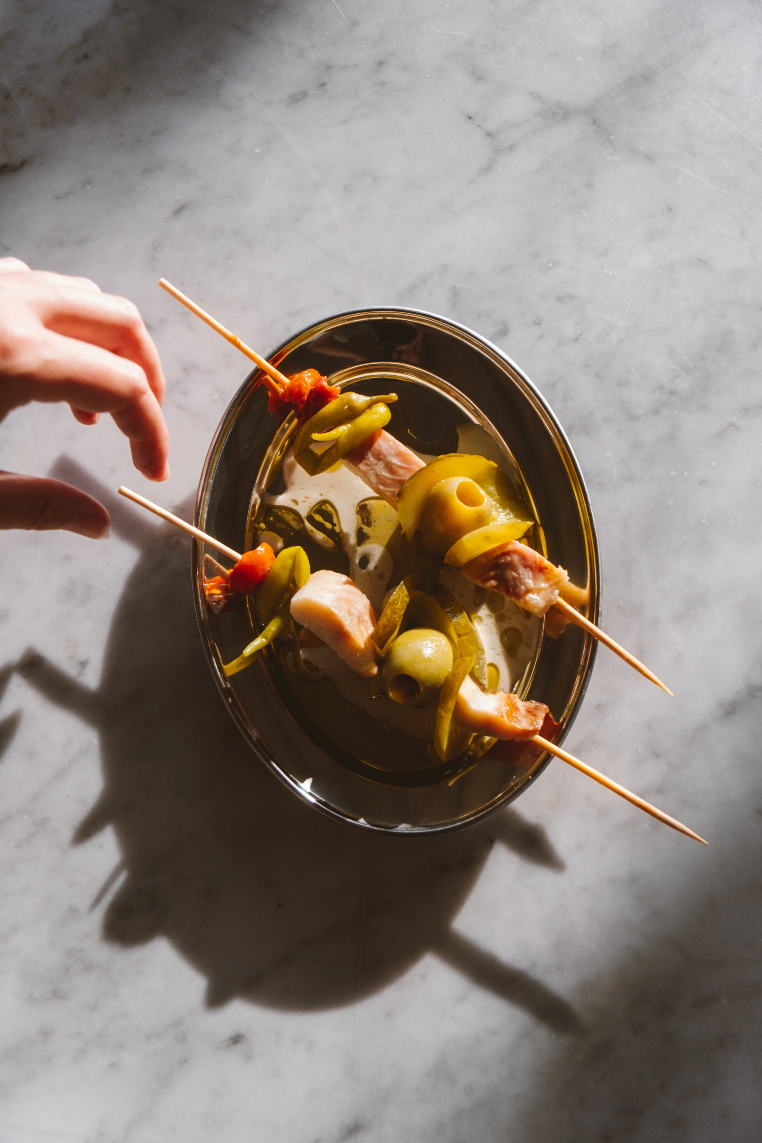 Mano colocando una brocheta con pollo, pimientos verdes y tomates cherry en un plato de metal con salsa, sobre una mesa de mármol.