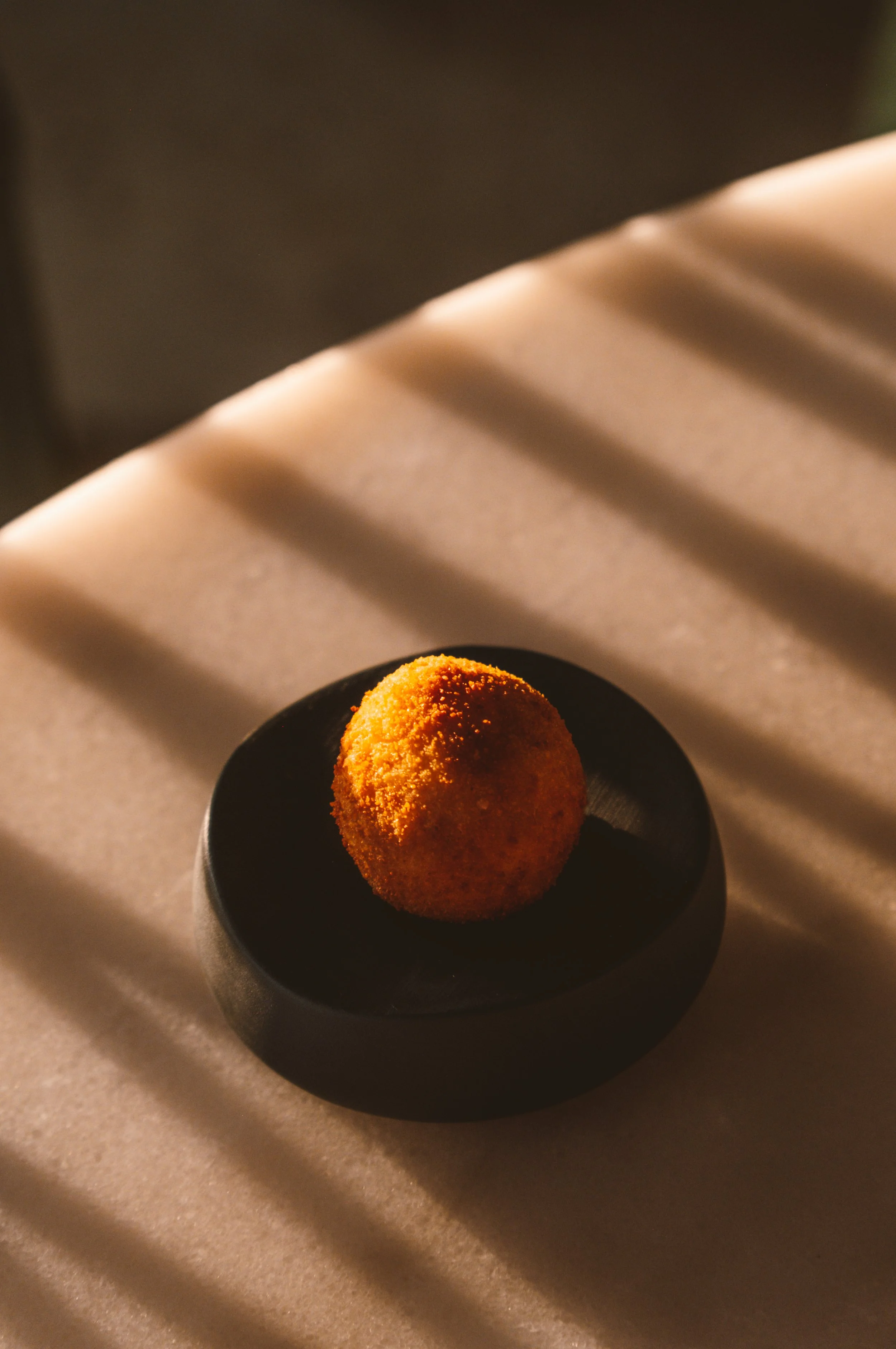 Una croqueta dorada en un plato negro sobre una superficie de color beige con sombras suaves.