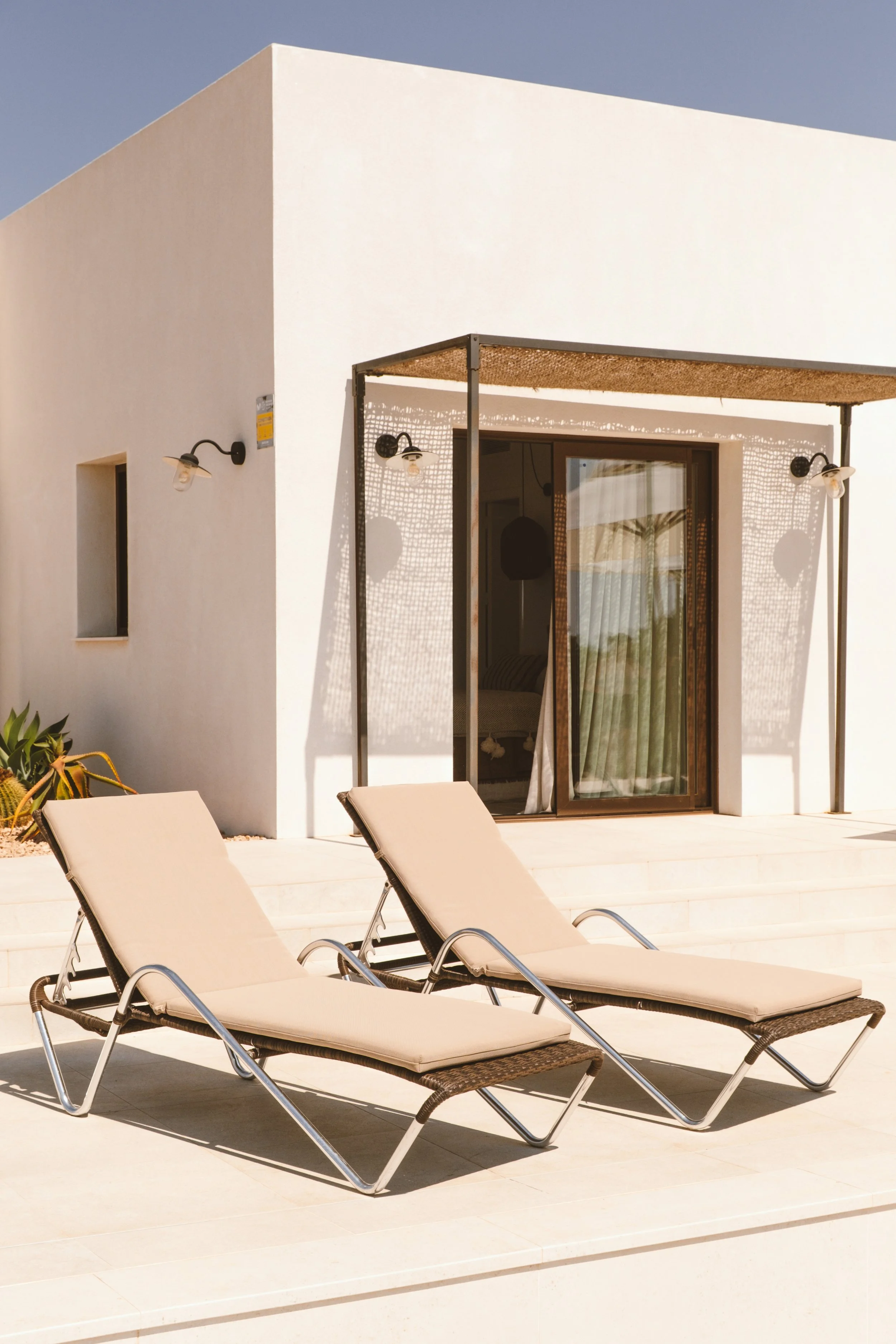 Sillas de descanso junto a una piscina con estructura de sombra en una casa moderna con paredes blancas y lámparas en el exterior.