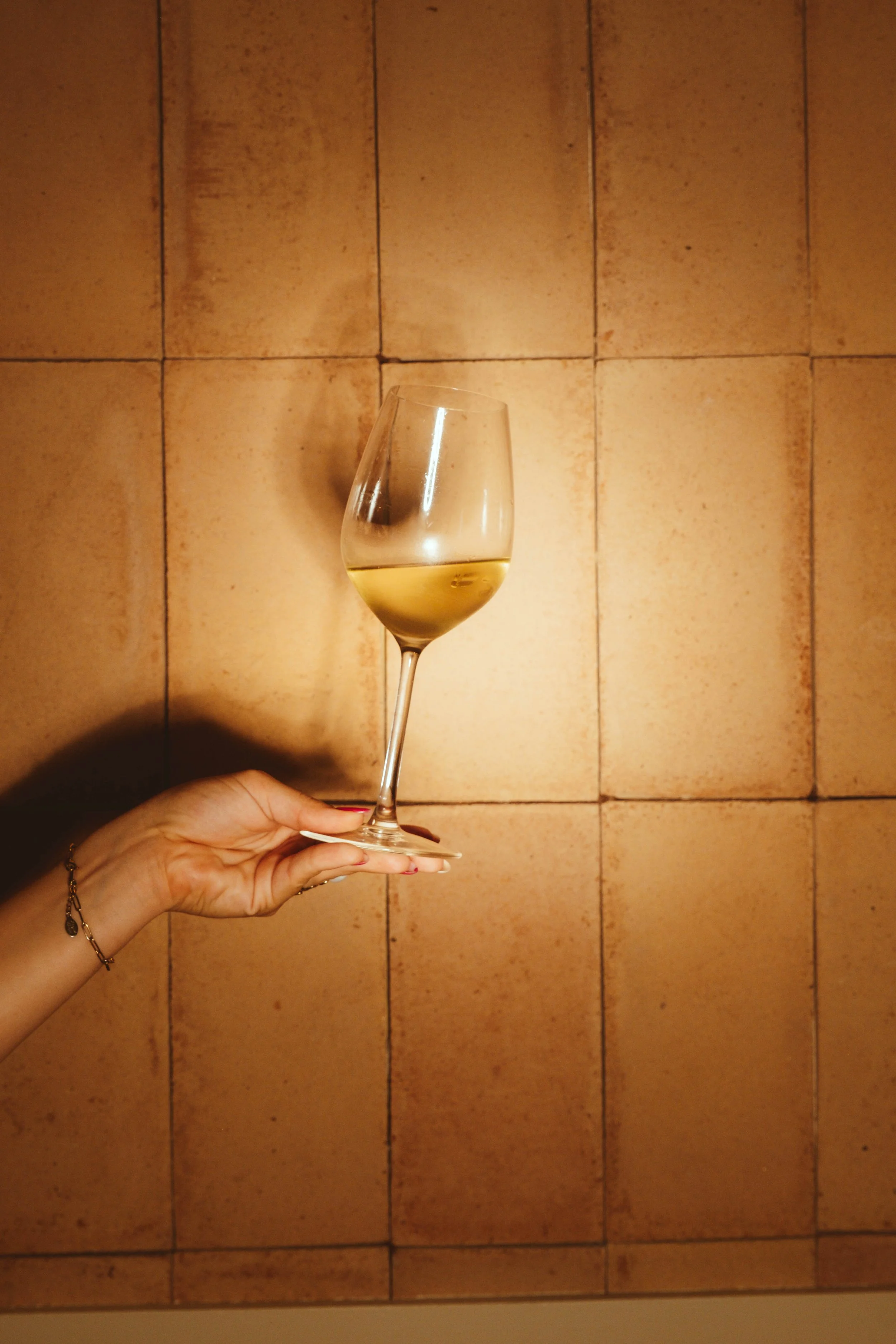 Una mano sostiene una copa de vino blanco en un ambiente con pared de azulejos marrones.