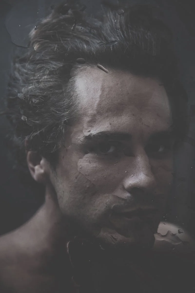 Retrato artístico de una persona con rostro cubierto en parte de barro, con expresión serena, rodeada de agua.