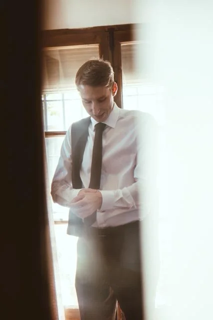 Joven hombre con camisa blanca y corbata, mirando su teléfono en una habitación con ventanas y cortinas de madera.