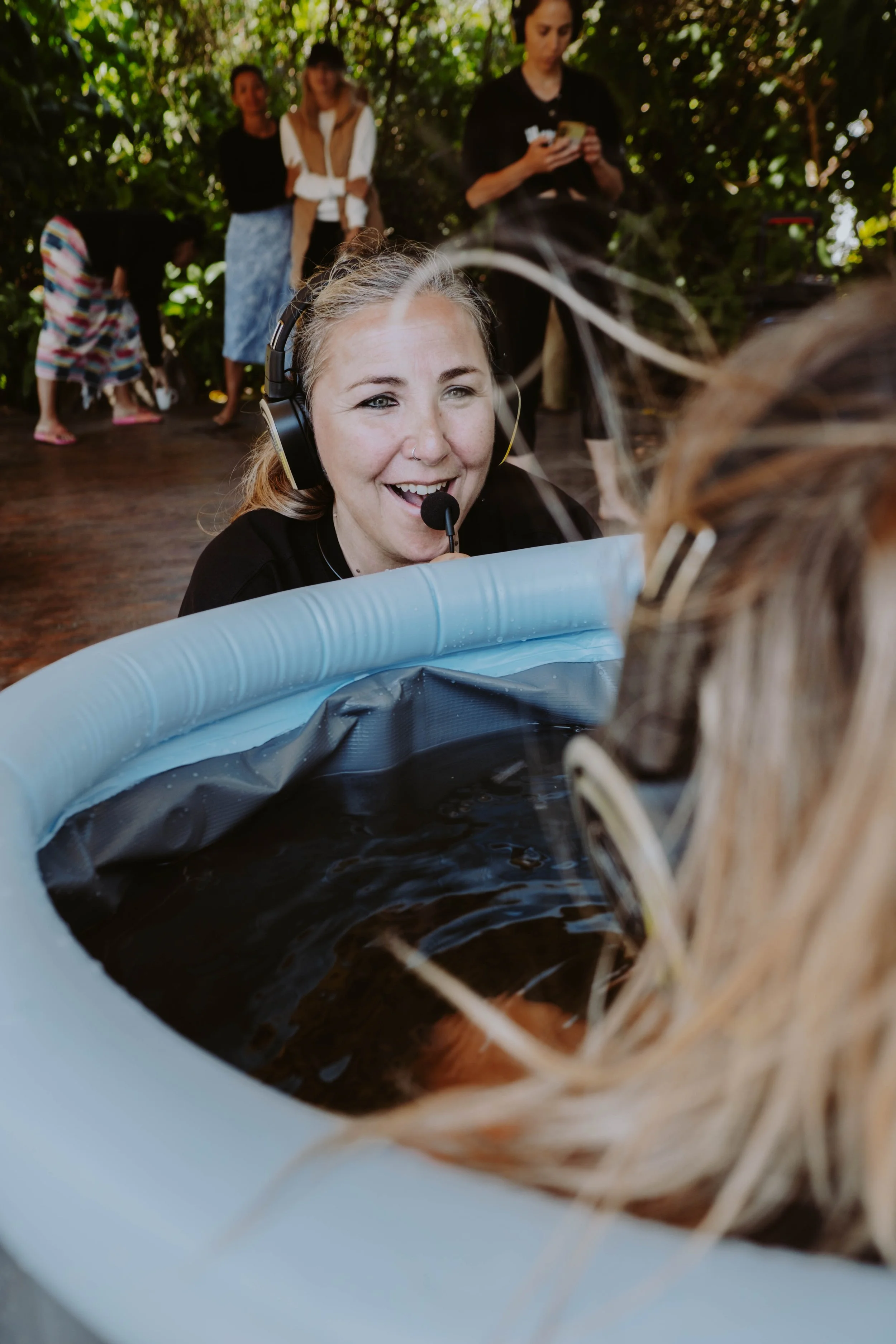 Mujer con auriculares y micrófono sonriendo mientras observa a un animal en una piscina inflable, en un entorno natural con árboles y más personas en el fondo.