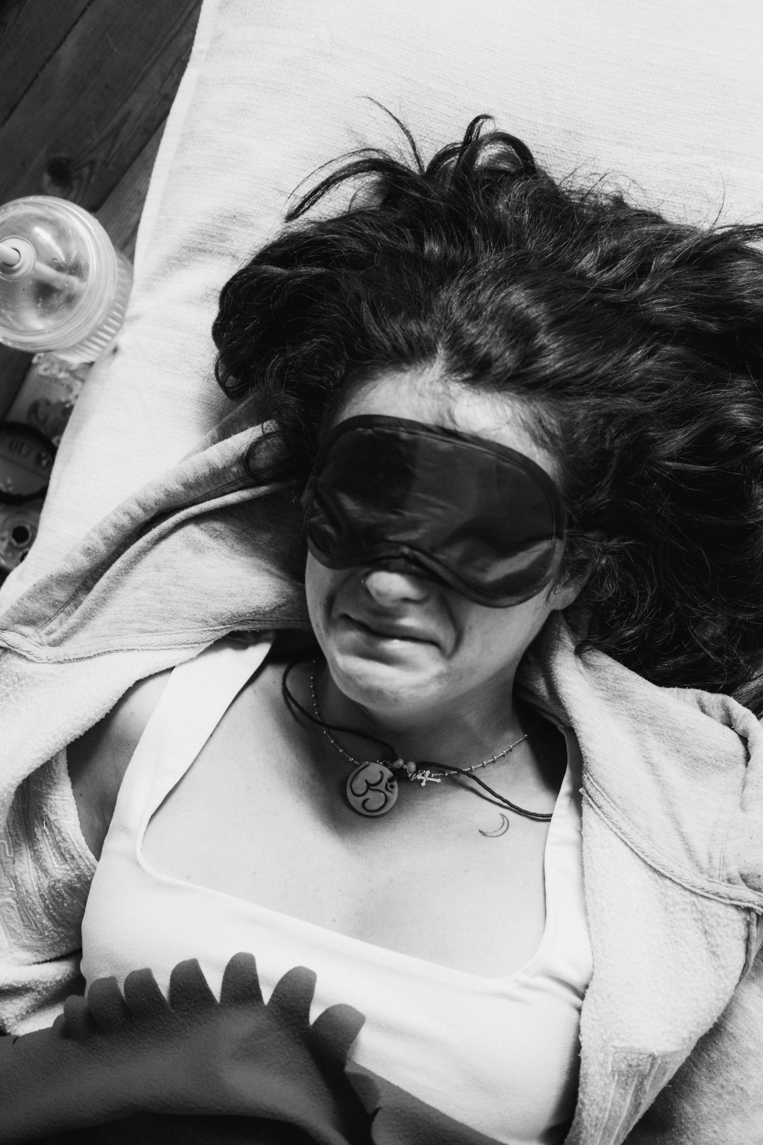 Mujer con cabello rizado oscuro y usando gafas de dormir oscuras, descansando sobre una cama con una manta y sosteniendo una botella de agua.