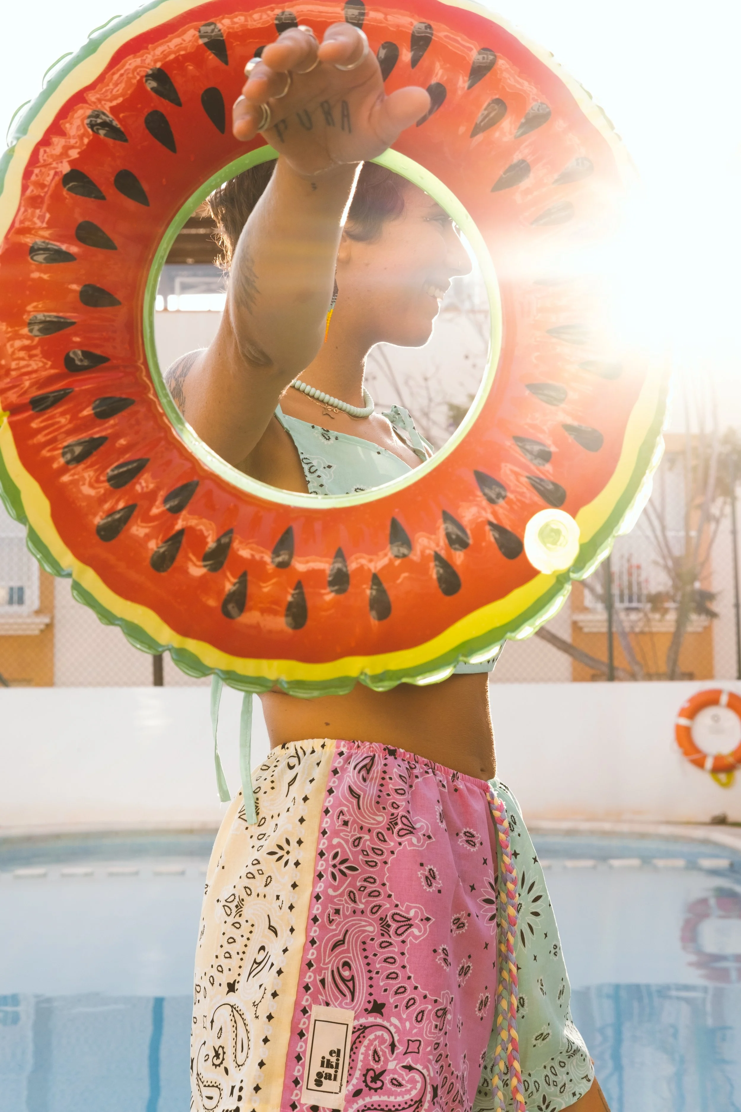 Persona sonriendo con gorro inflable de sandía, junto a la piscina, al atardecer, viste ropa colorida y accesorios de este estilo.