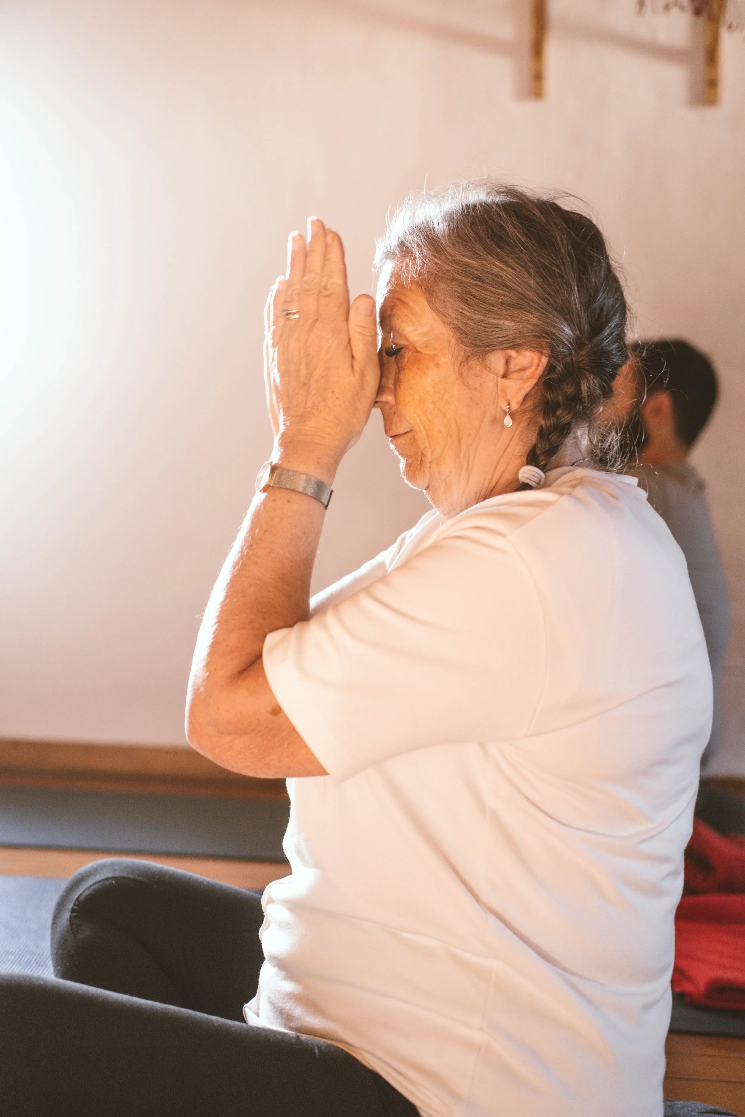 Anciana con cabello gris y trenza, de perfil, en postura de oración, usando camiseta blanca y pendientes colgantes, participando en actividad espiritual en interior con fondo difuso.