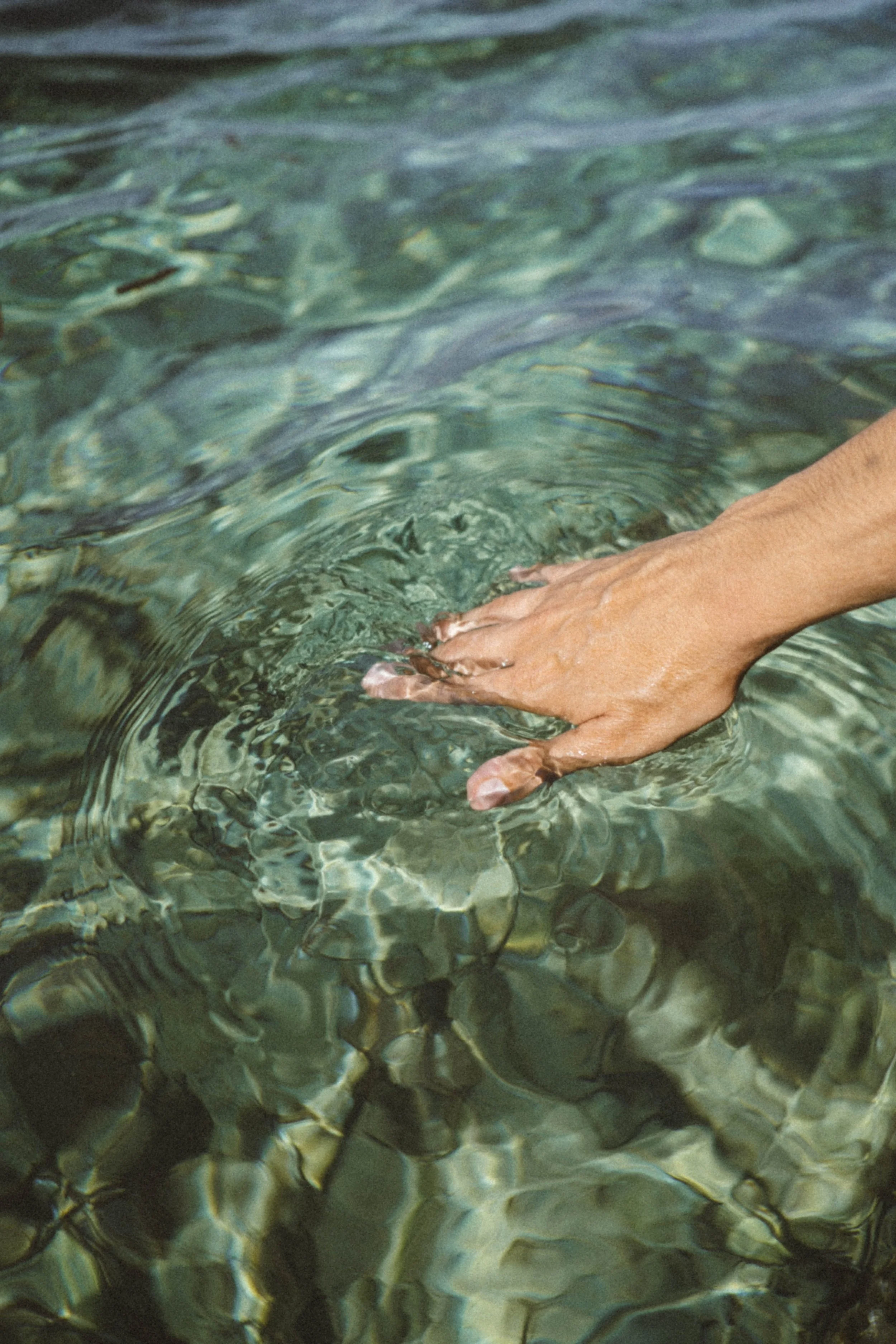 Persona sumergiendo los pies en agua transparente, que refleja el fondo de piedras en el río o mar.