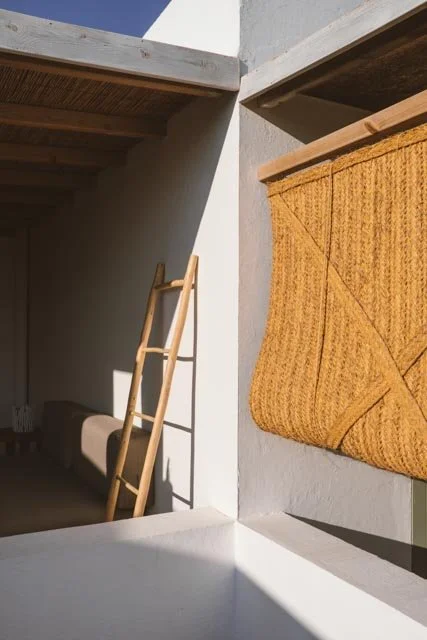Un espacio exterior con paredes blancas, una escalera de madera apoyada contra la pared y una cortina de fibra natural en la ventana