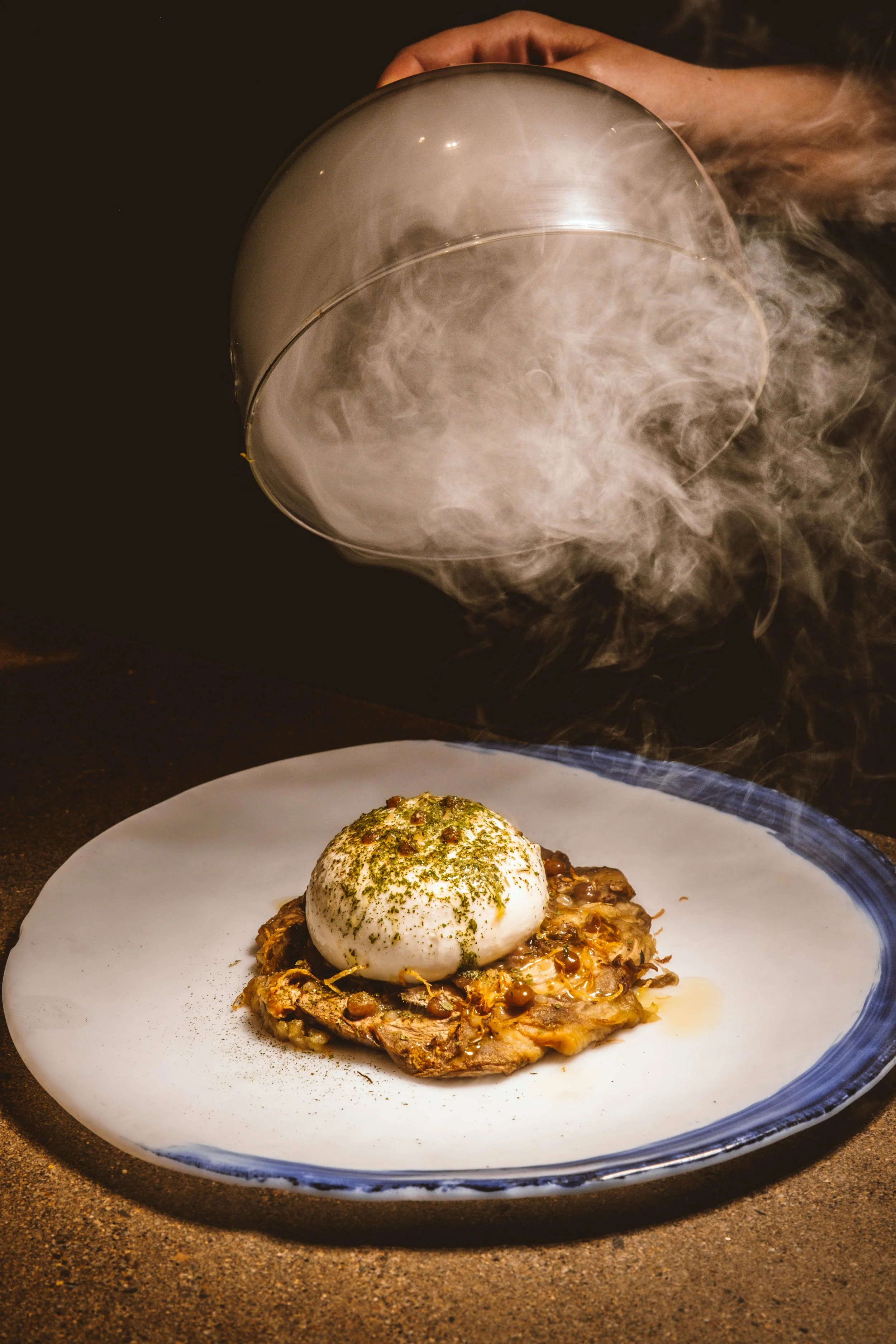 Plato con huevo pochado, hierbas y acompañamiento de carne en un plato blanco con borde azul. Vapores y niebla de hielo seco sobre el plato.