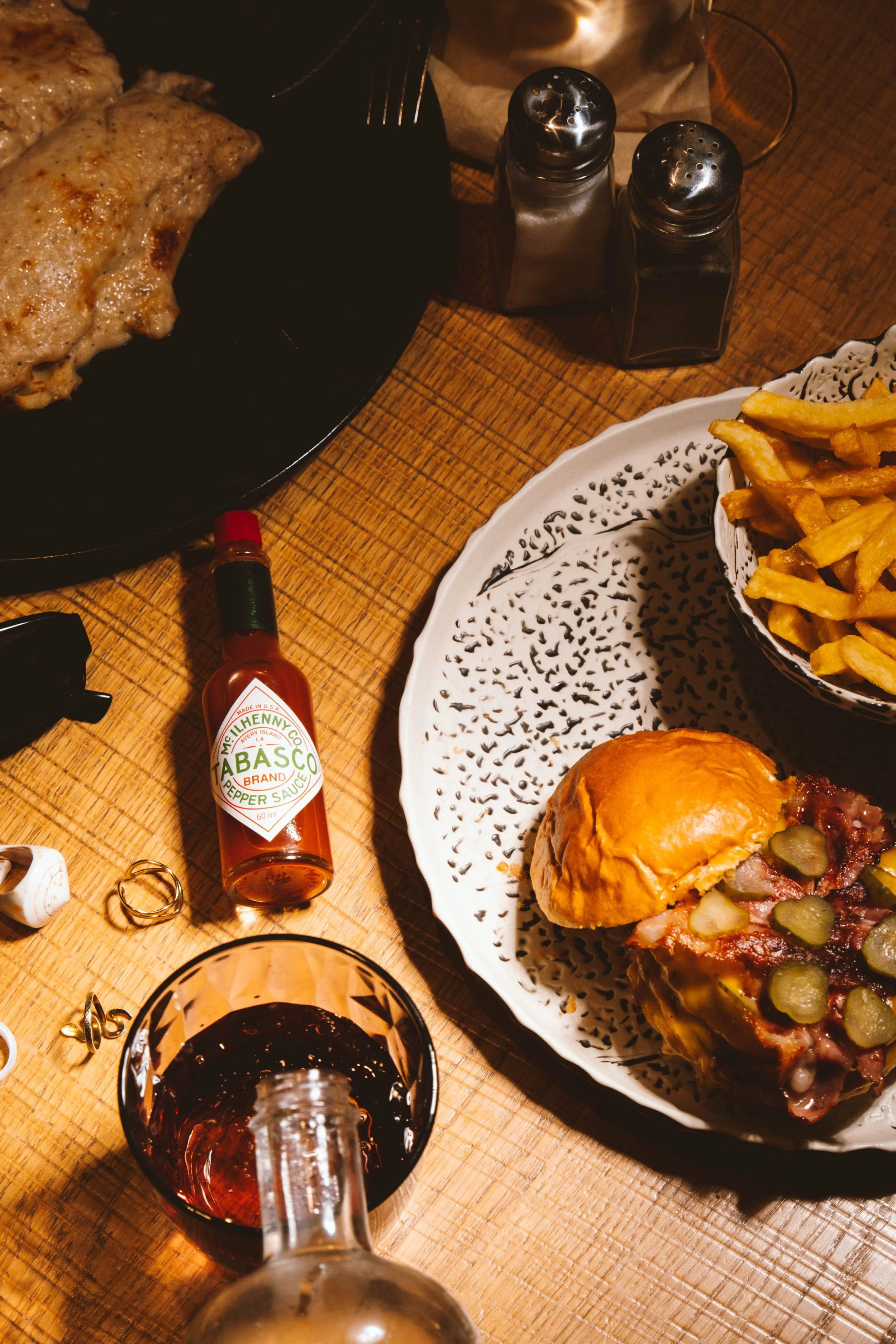 Plato de un sándwich de carne con pepinillos, papas fritas, salsa picante, salero, pimienta, salsa Tabasco, y una bebida roja en un vaso.