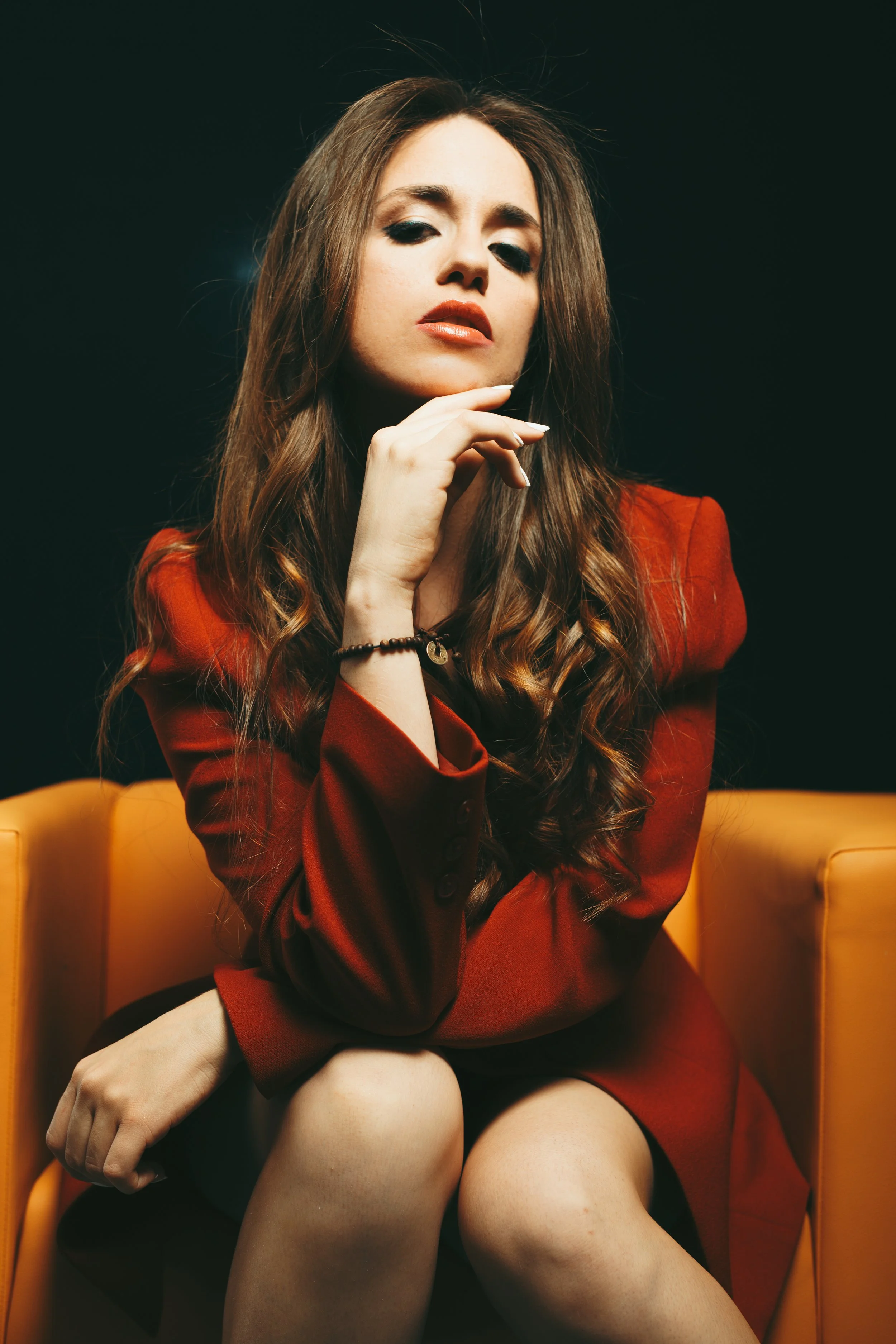 Mujer con cabello rizado, vestida con saco rojo, en una silla amarilla, mirando hacia la cámara con expresión seria, en un fondo negro.