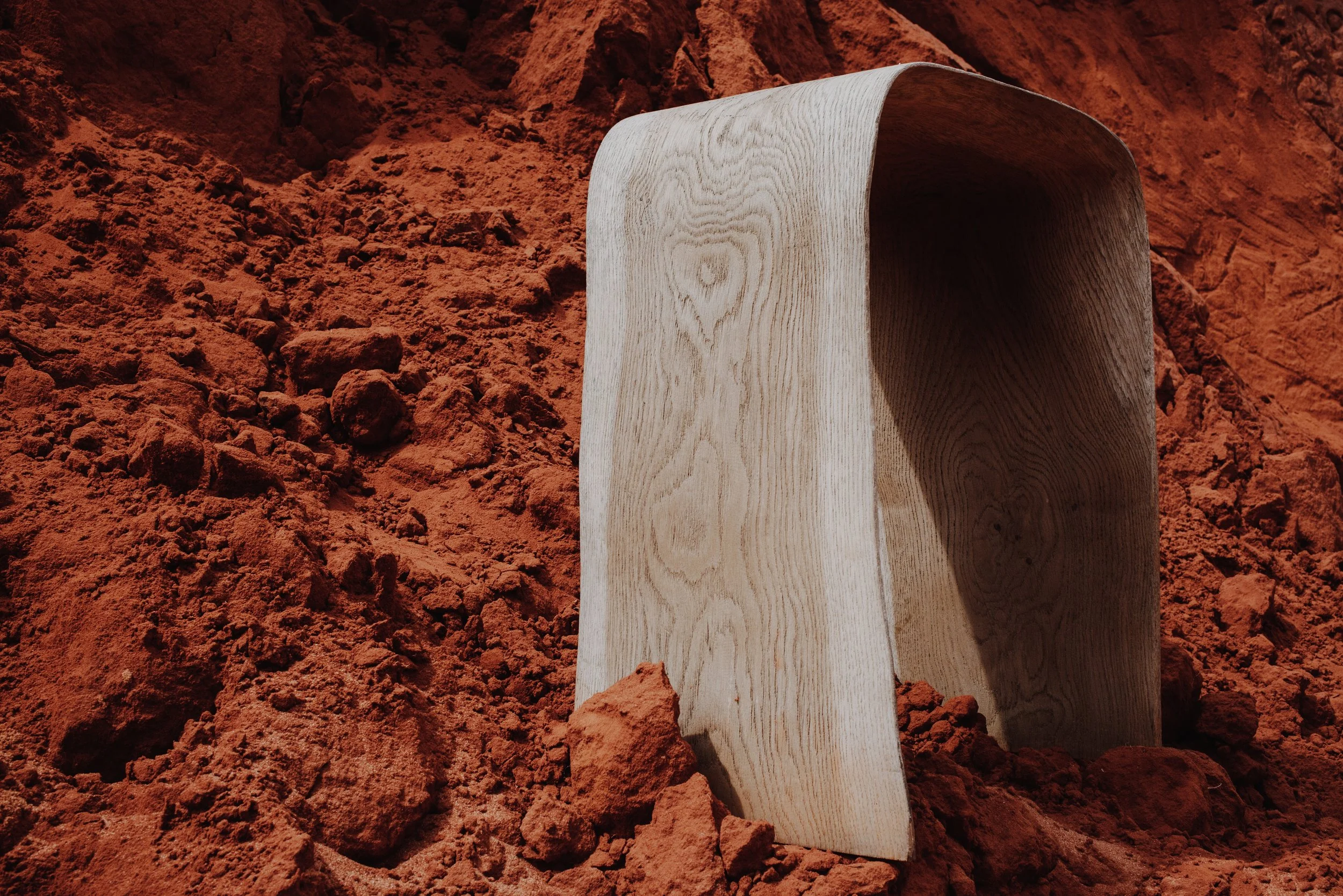 Un trozo de madera clara, recto y con vetas visibles, está parcialmente enterrado en un suelo de tierra roja y seca.