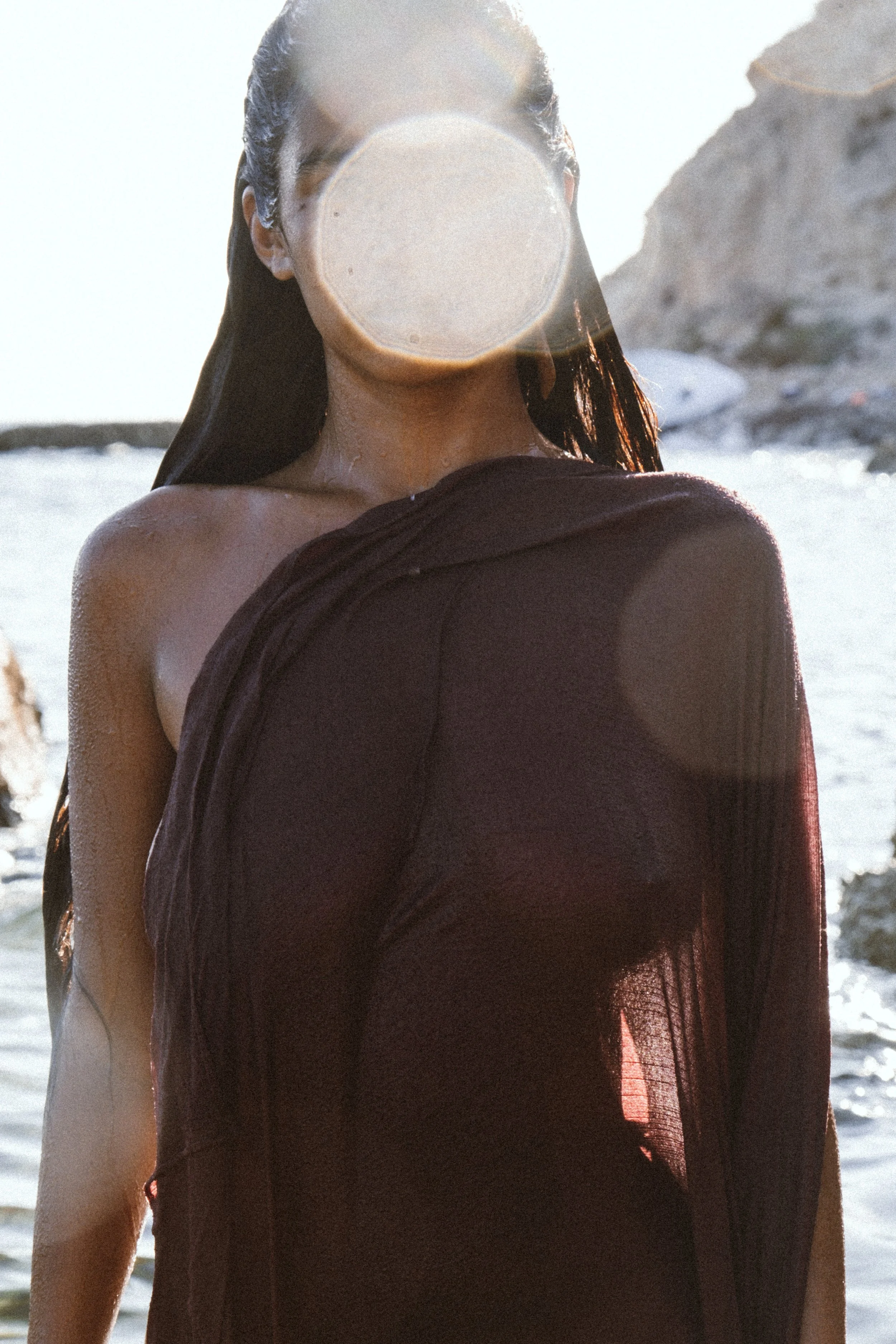 Mujer con cabello largo y suelto al frente, vestida con una prenda oscura, de espaldas, junto a un cuerpo de agua y rocas, con luz solar brillante.