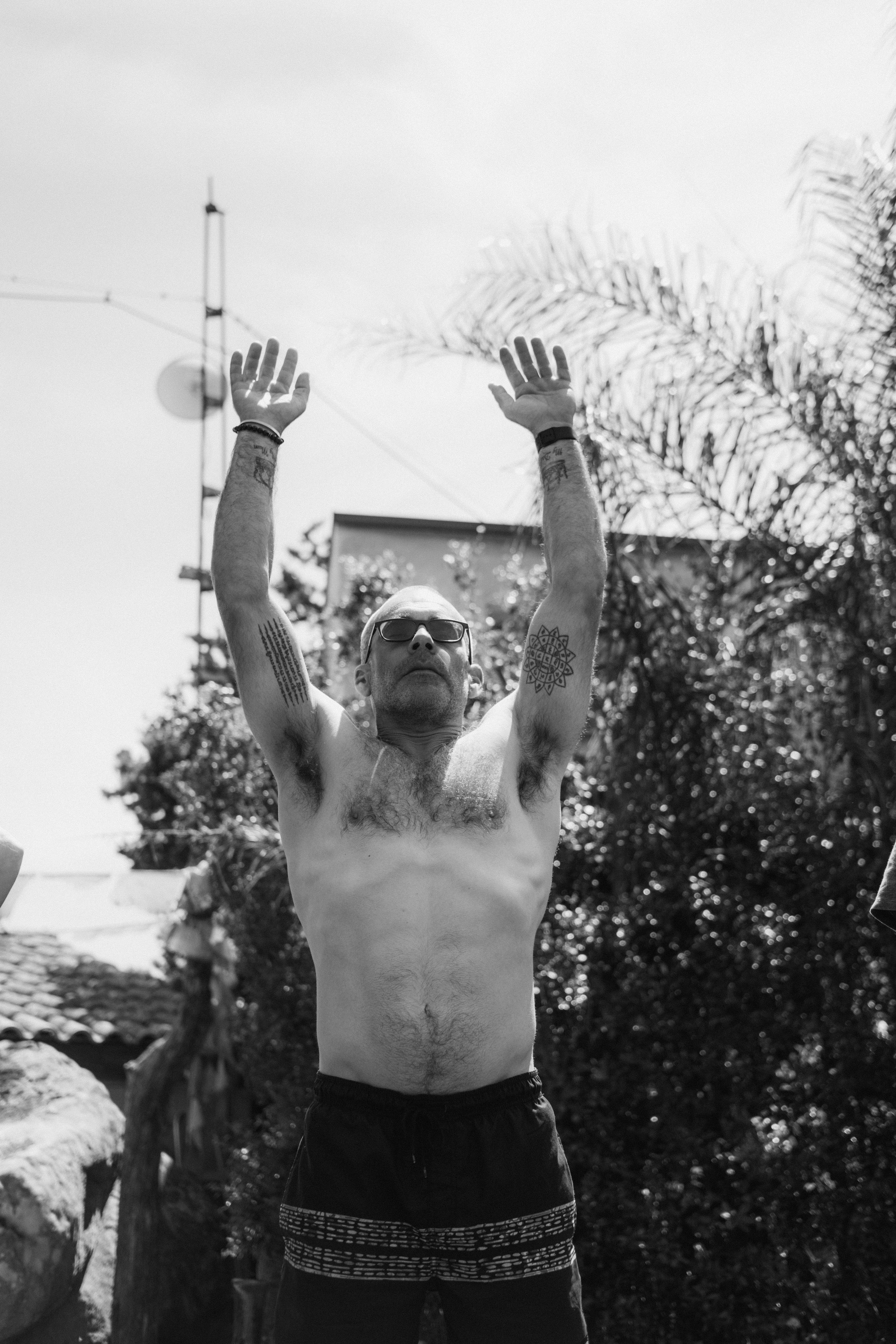 Hombre sin camiseta con gafas levantando los brazos, tatuajes en los brazos, en un entorno exterior con árboles y cables eléctricos.