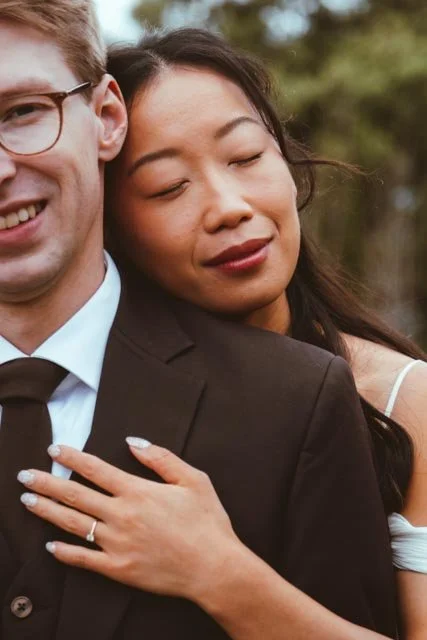 Una pareja abraza con alegría, ella con los ojos cerrados y sonrisa tranquila, él parcialmente visible con gafas y vestido formal, en un entorno al aire libre.