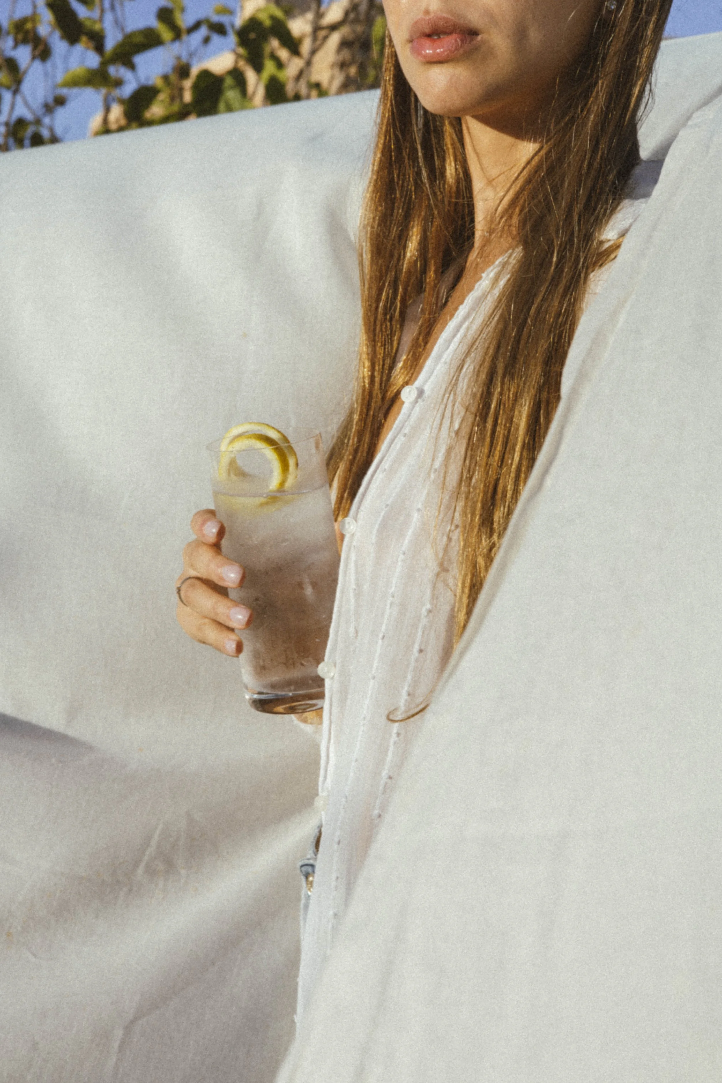 Mujer con cabello largo y rubio, usando blusa blanca, sosteniendo un vaso con bebida y rodaja de limón, en un entorno con fondo de árboles y cielo azul.