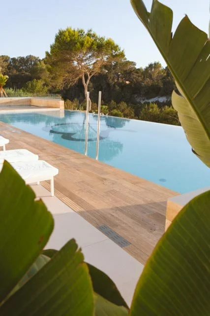 Piscina al aire libre con agua azul, rodeada de palmas tropicales y árboles, en una zona de descanso con tumbonas blancas.