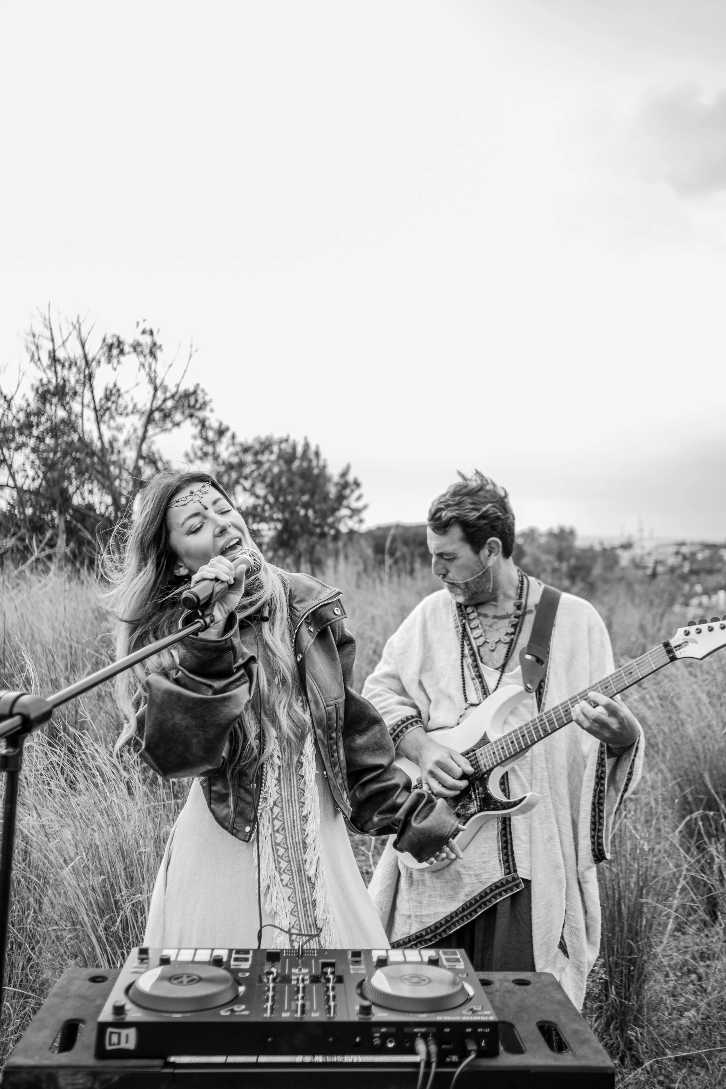 Un grupo de dos personas tocando música en un campo abierto. La mujer canta y toca la guitarra, mientras que el hombre también toca la guitarra. En primer plano, una mesa con equipo de DJ.