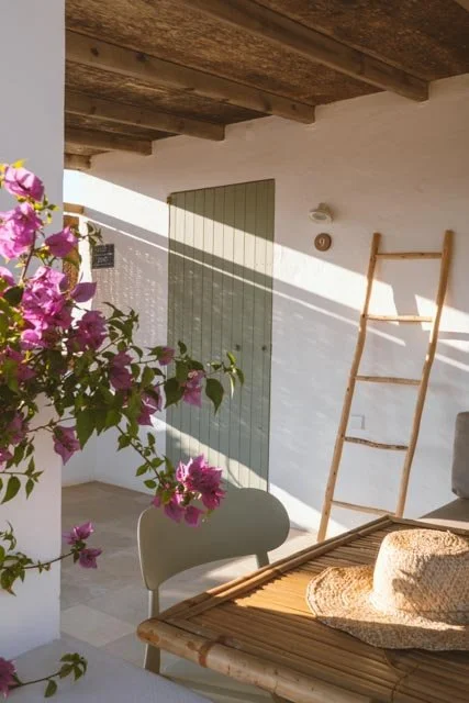 Interior de una terraza con pared blanca, una puerta de madera, plantas con flores moradas, una silla y un sombrero de paja sobre una mesa de madera.