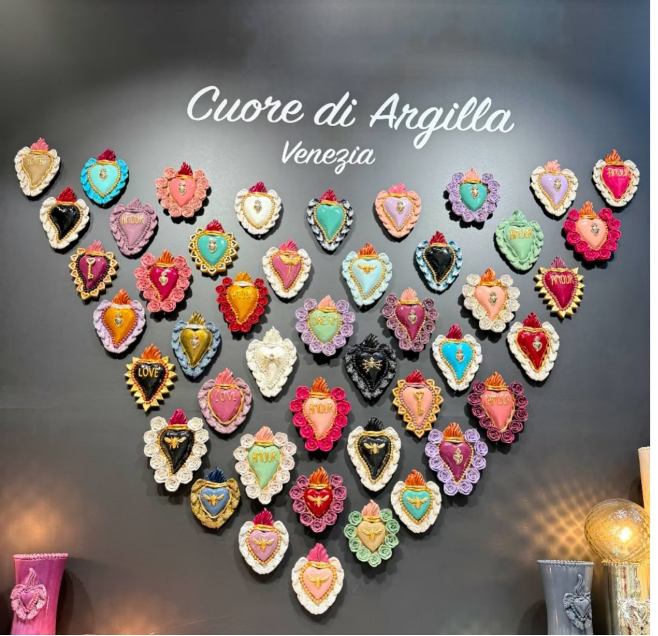 Collection de hearts en céramique colorés, décorés de fleurs en pâte à sucre, avec le mot « amour » écrit dessus, exposés sur une surface grise. Titre en français : « Coeur di Argilla Venezia ».