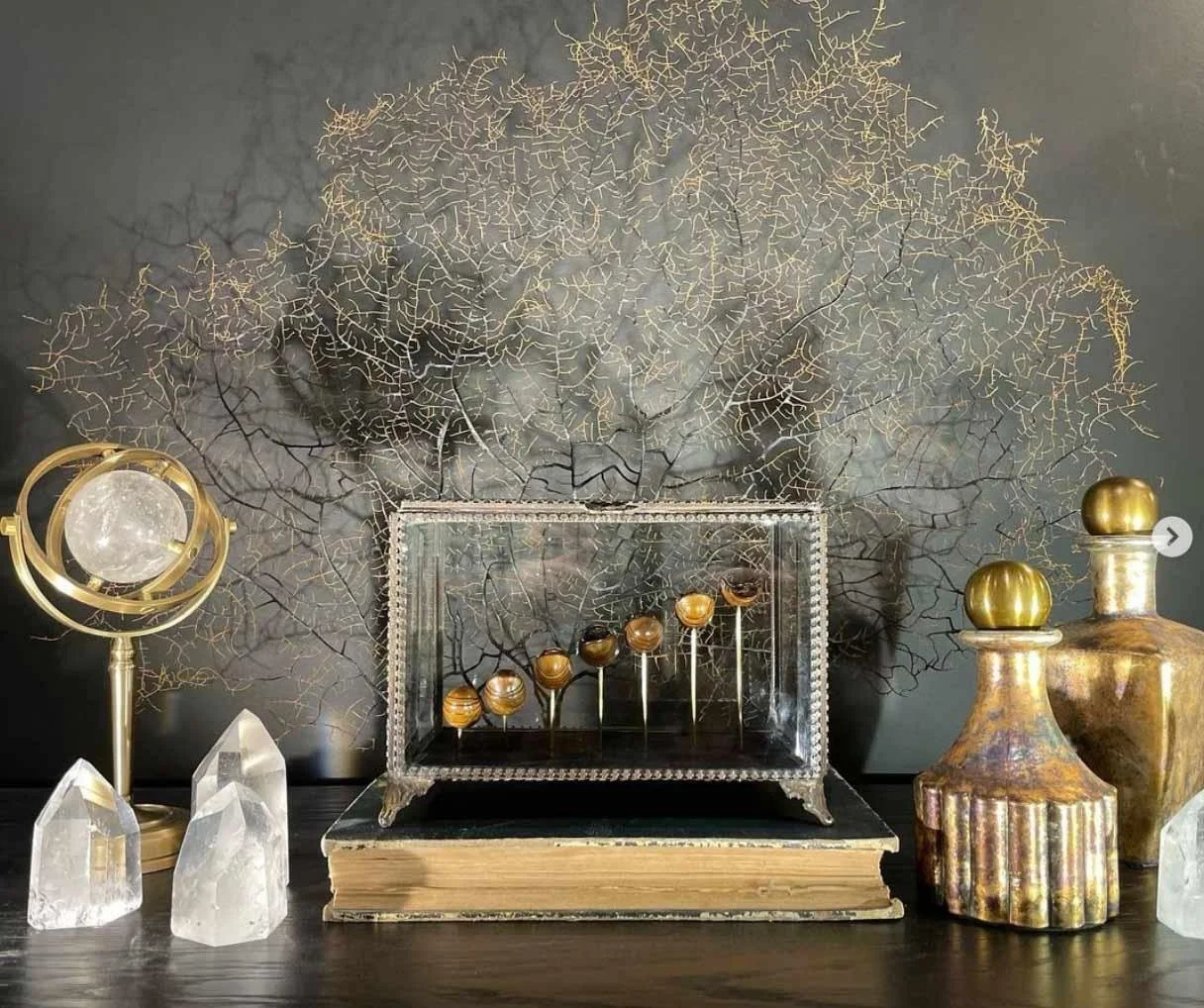 Décoration artisanale avec un fond en branches d'arbre dorées, une boîte en verre avec des billes en bois, deux vases ancien avec des sphères dorées, des cristaux et un livre en dessous.