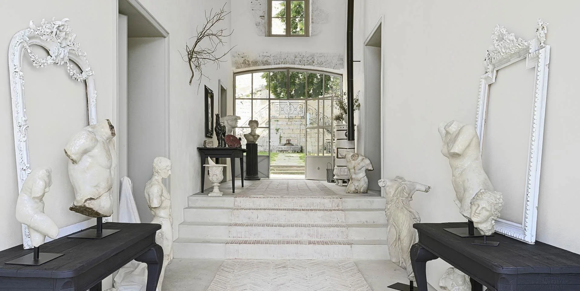 Intérieur d'une galerie d'art avec sculptures en marbre de bustes et corps, cadres vides ornés de sculptures et décoration blanche, vue vers une porte-fenêtre donnant sur un jardin.