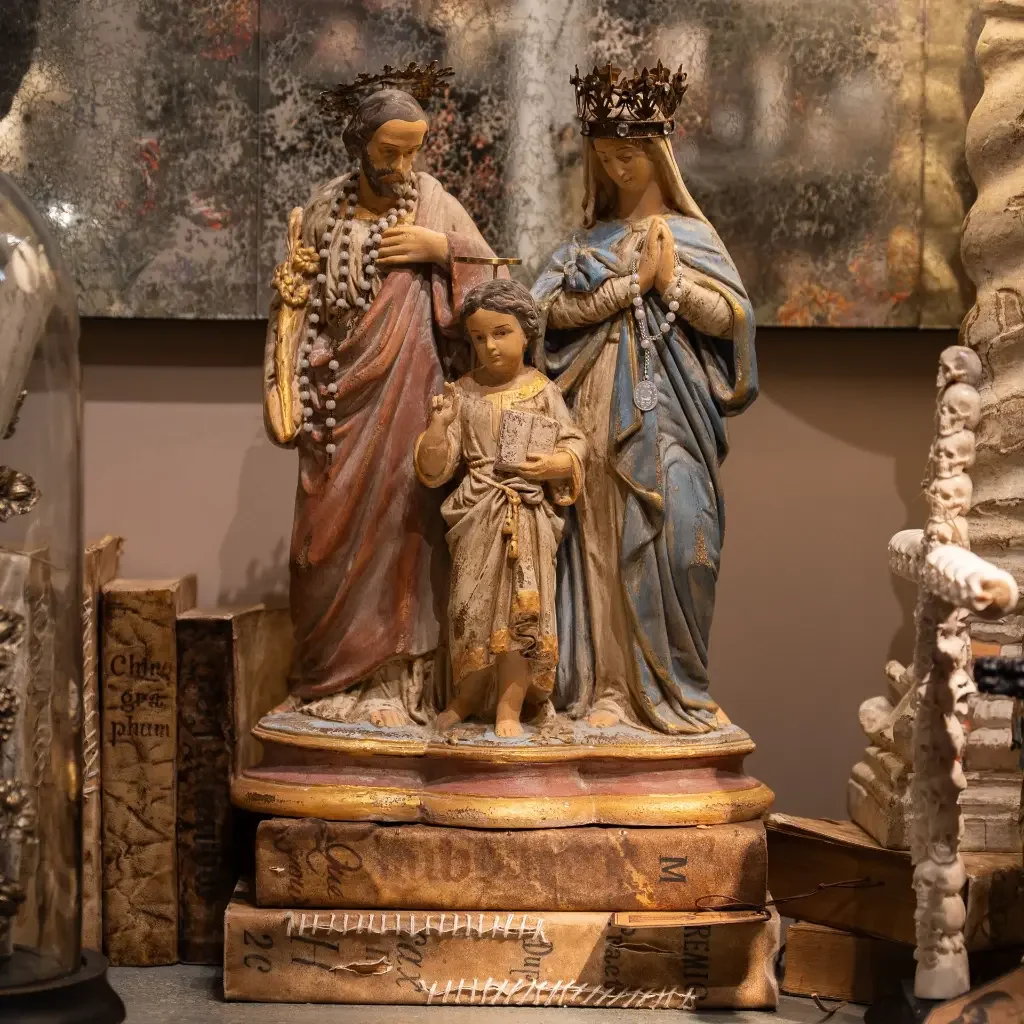 Une sculpture religieuse représentant la Sainte Famille avec Jésus, Marie et Joseph, portant des couronnes et des chapelets, placée sur des livres anciens.