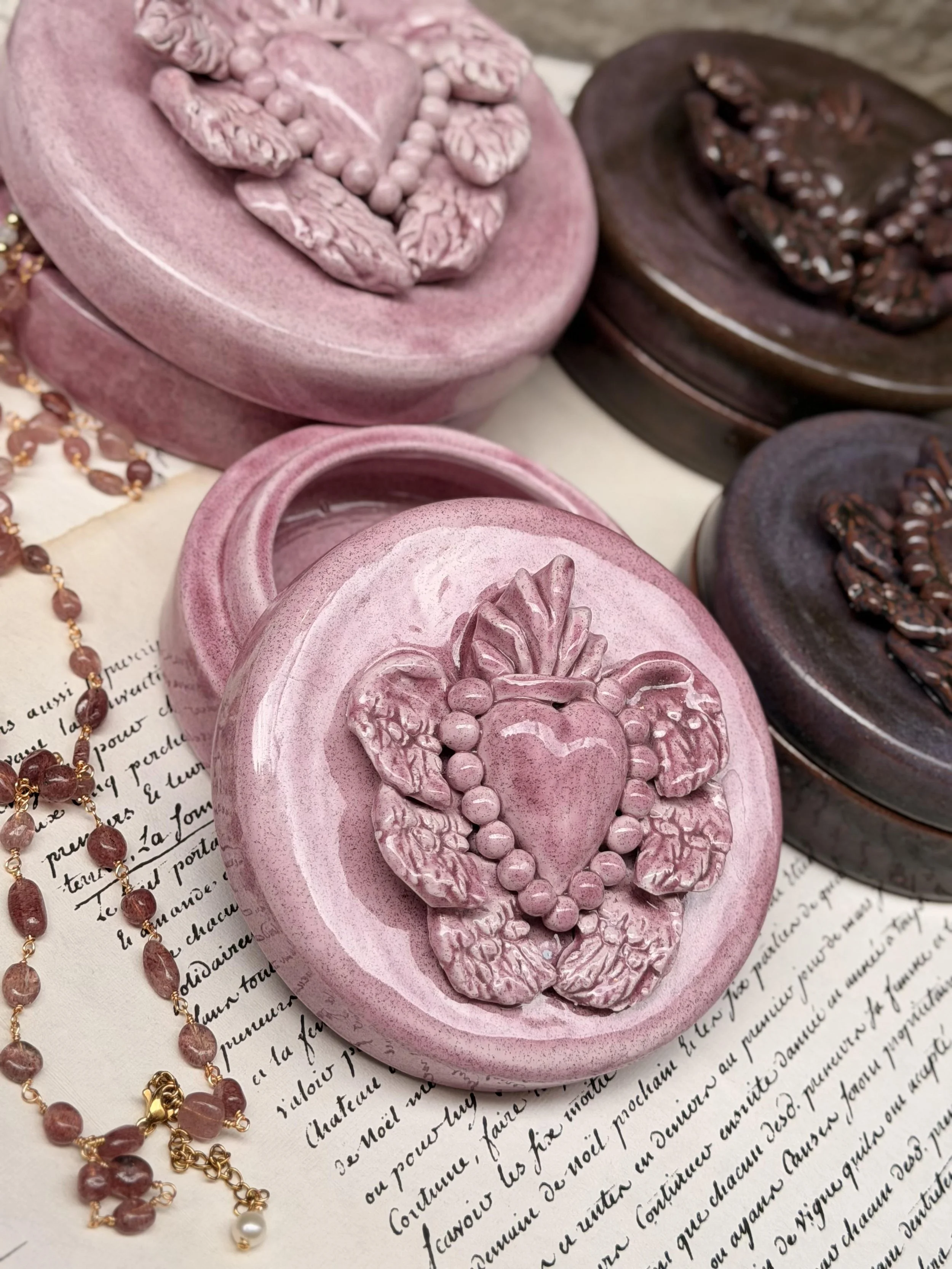 Deux boîtes de savons en forme de cœurs, l'une rose avec un motif floral et l'autre marron avec un motif différent, posées sur un fond de papier avec du texte manuscrit. À côté, un collier de perles en forme de petits cœurs marron et une perle blanche, laissant entrevoir une ambiance élégante et décorative.