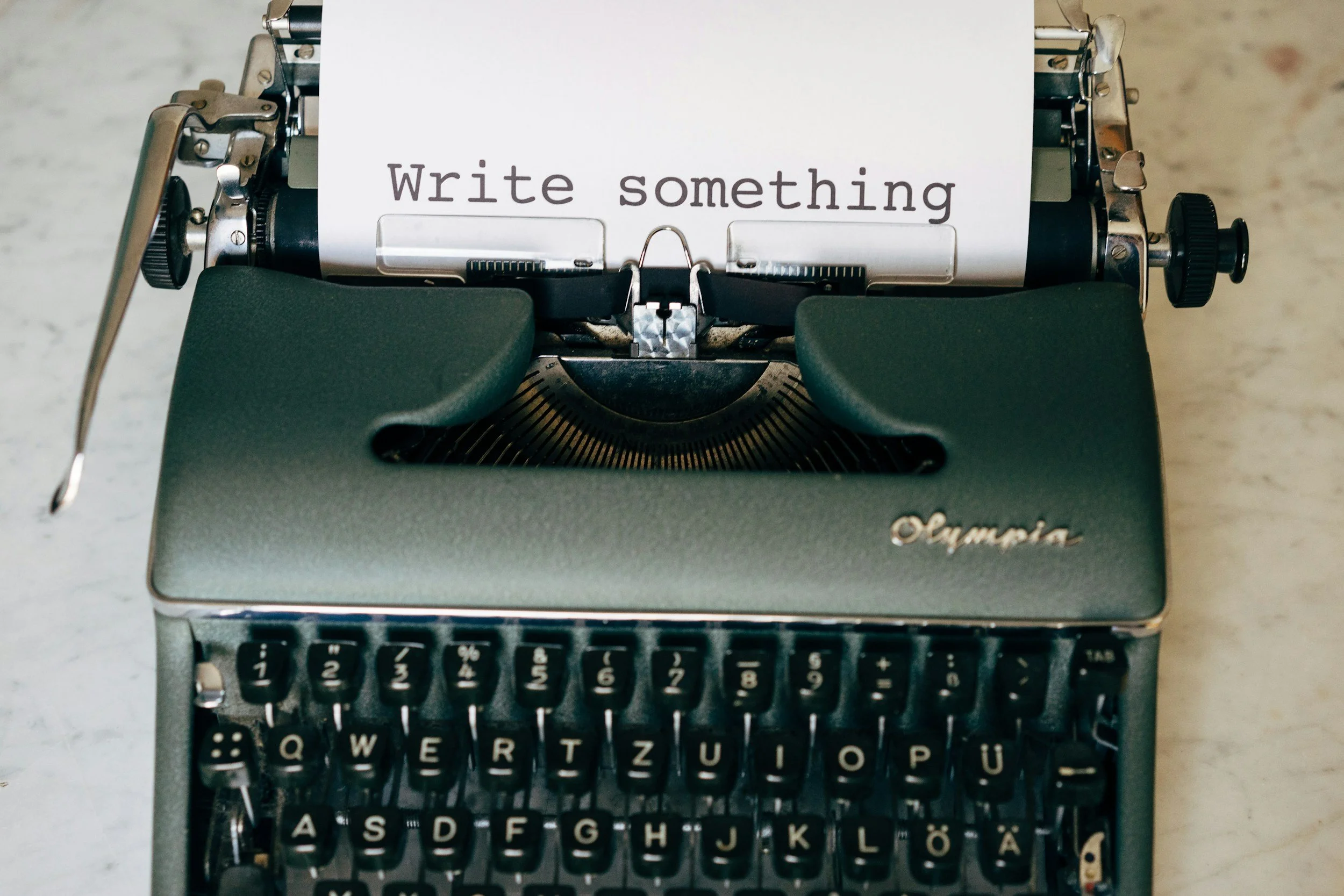 A vintage typewriter with a piece of paper inserted, displaying the words 'Write something'. The typewriter has black keys with white lettering and metal components, resting on a light-colored surface.