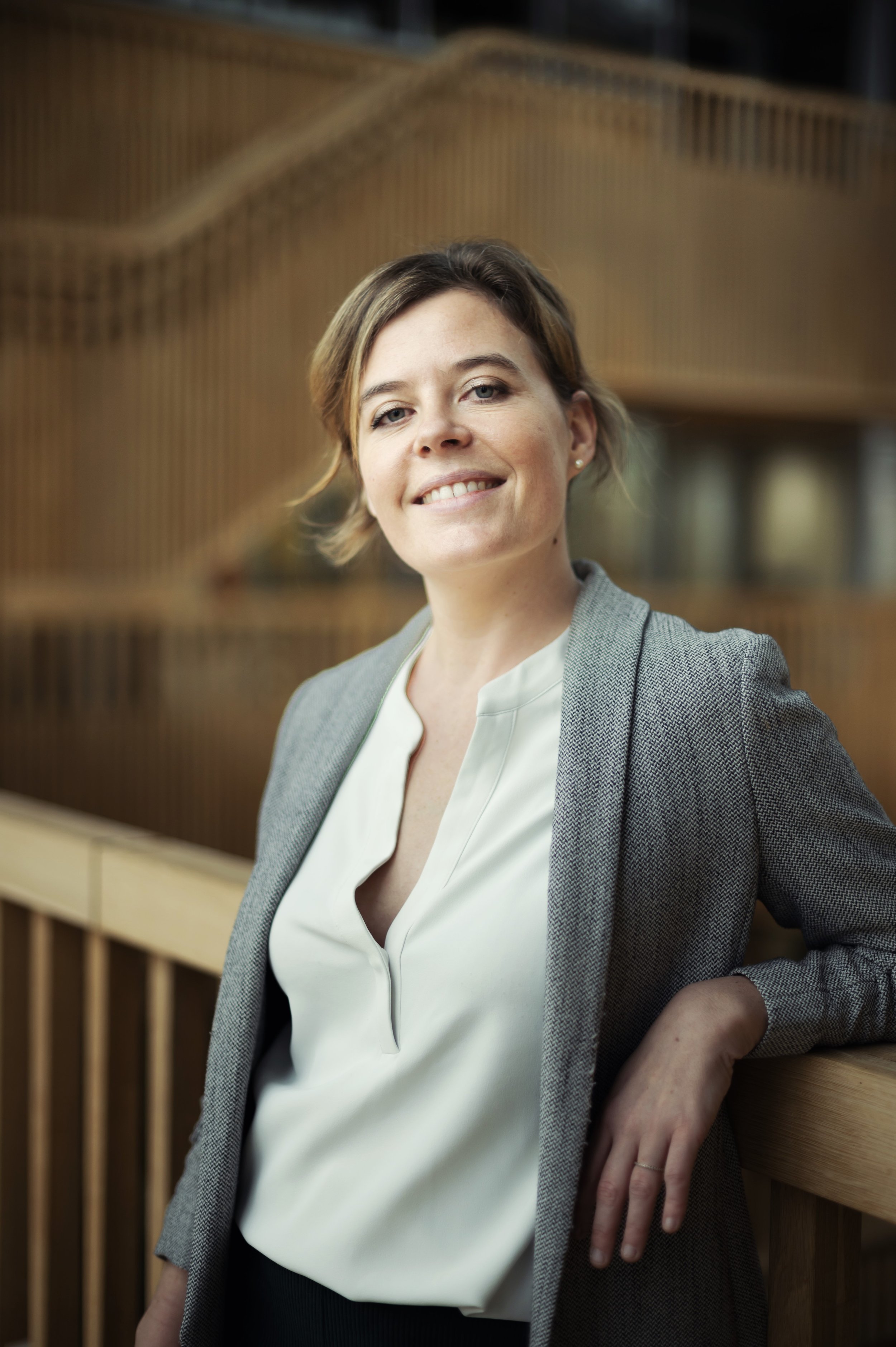 A woman with short brown hair and light skin is smiling and leaning against a wooden railing, wearing a gray blazer and a white blouse.