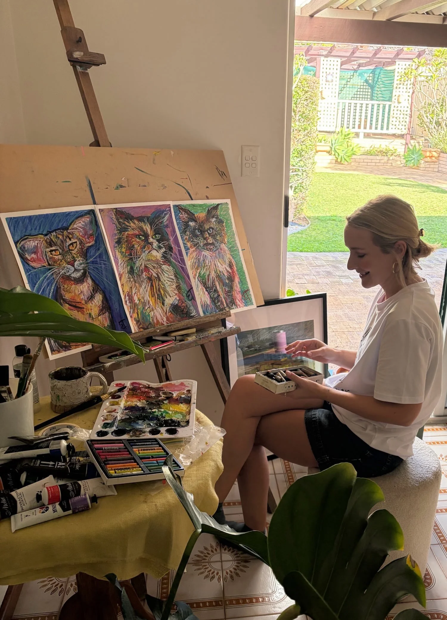 The female artist sat at her easel with art supplies, painting colorful portraits of cats on paper. Behind her is a large window showing a garden with green grass and plants.