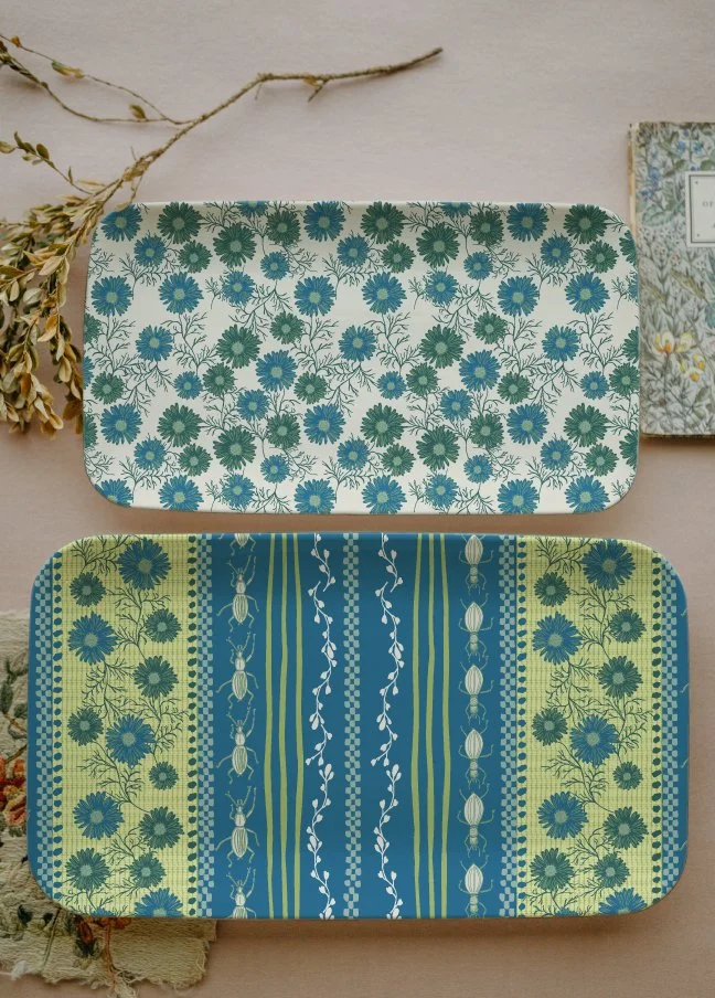 Two rectangular ceramic trays with floral and striped patterns, one above the other, on a neutral background with dried flowers and a calendar nearby.