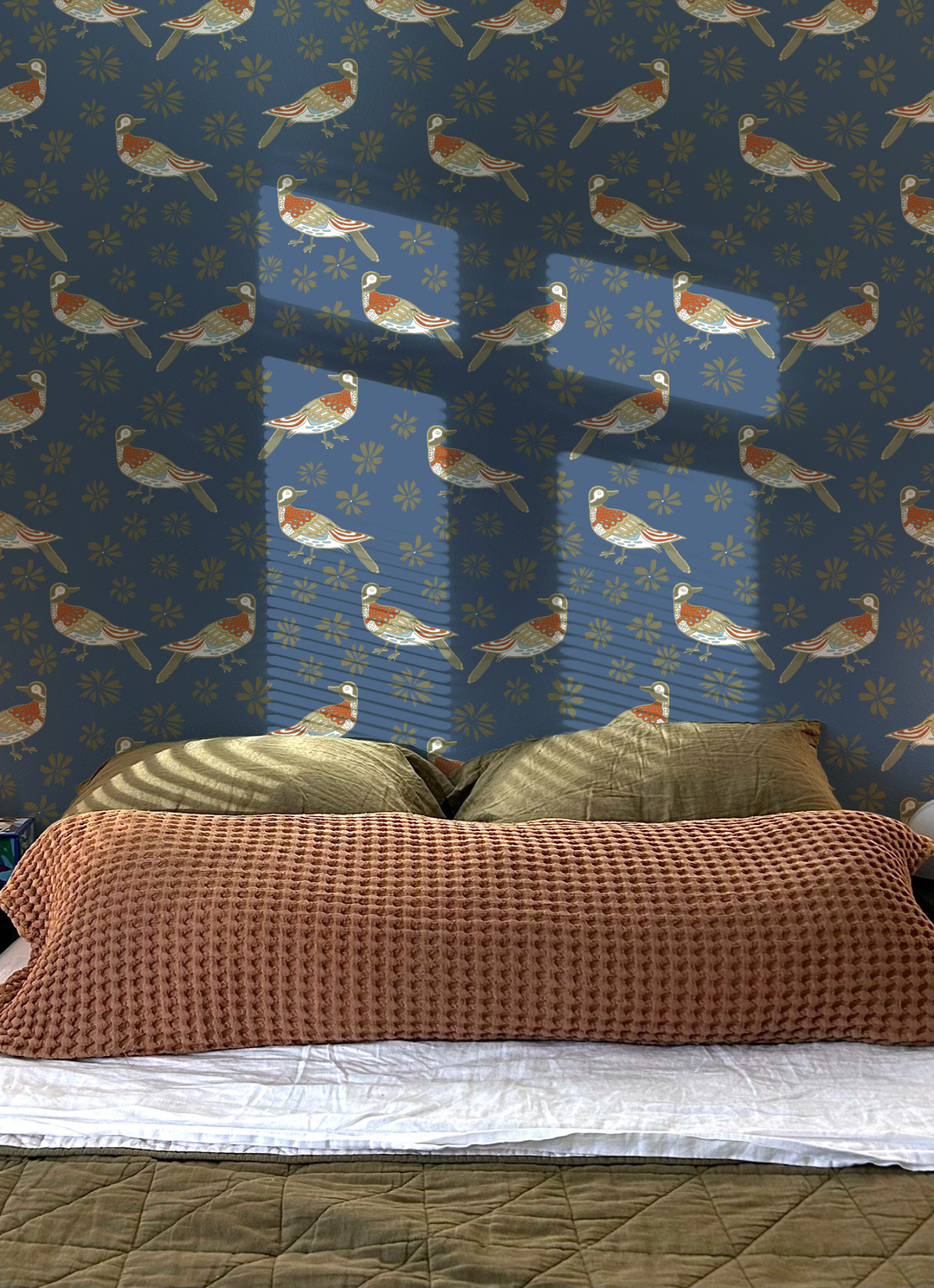 A bedroom with a bed, brown textured blanket, beige pillows, a pink checkered blanket, and a green quilted bedspread. The wall has a blue wallpaper with a pattern of birds and yellow flowers. Sunlight casts shadows on the wallpaper.