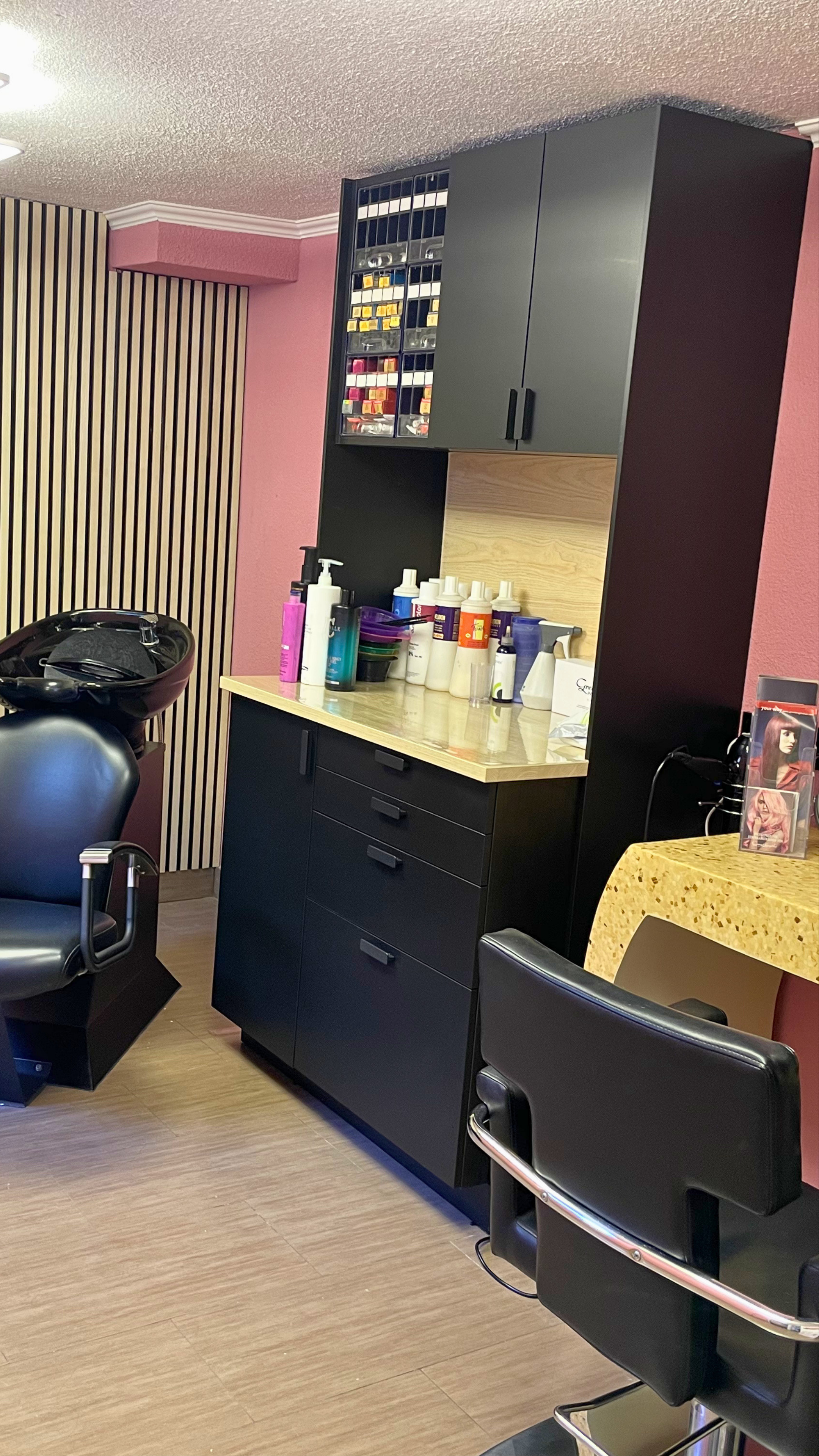 Friseursalon Salon Maya bei Hamburg mit schwarzen Stühlen, einer schwarzen Schubladenkommode, einer schwarzen Wand mit Tür und einer Ablage mit Pflegeprodukten. Im Hintergrund ein Spiegel und eine Wand mit Holzverkleidung.
