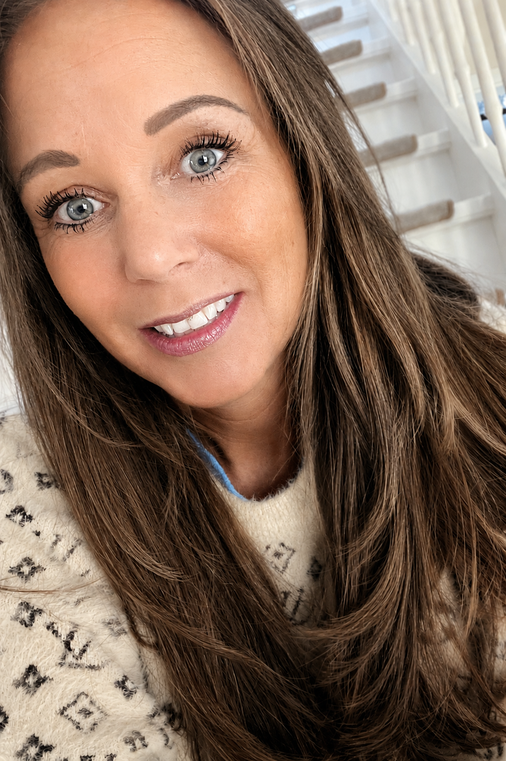 Nahaufnahme lachender Mandy Yazici mit langen braunen Haaren, blauen Augen und Make-up, vor einem weißen Hintergrund mit Treppen.